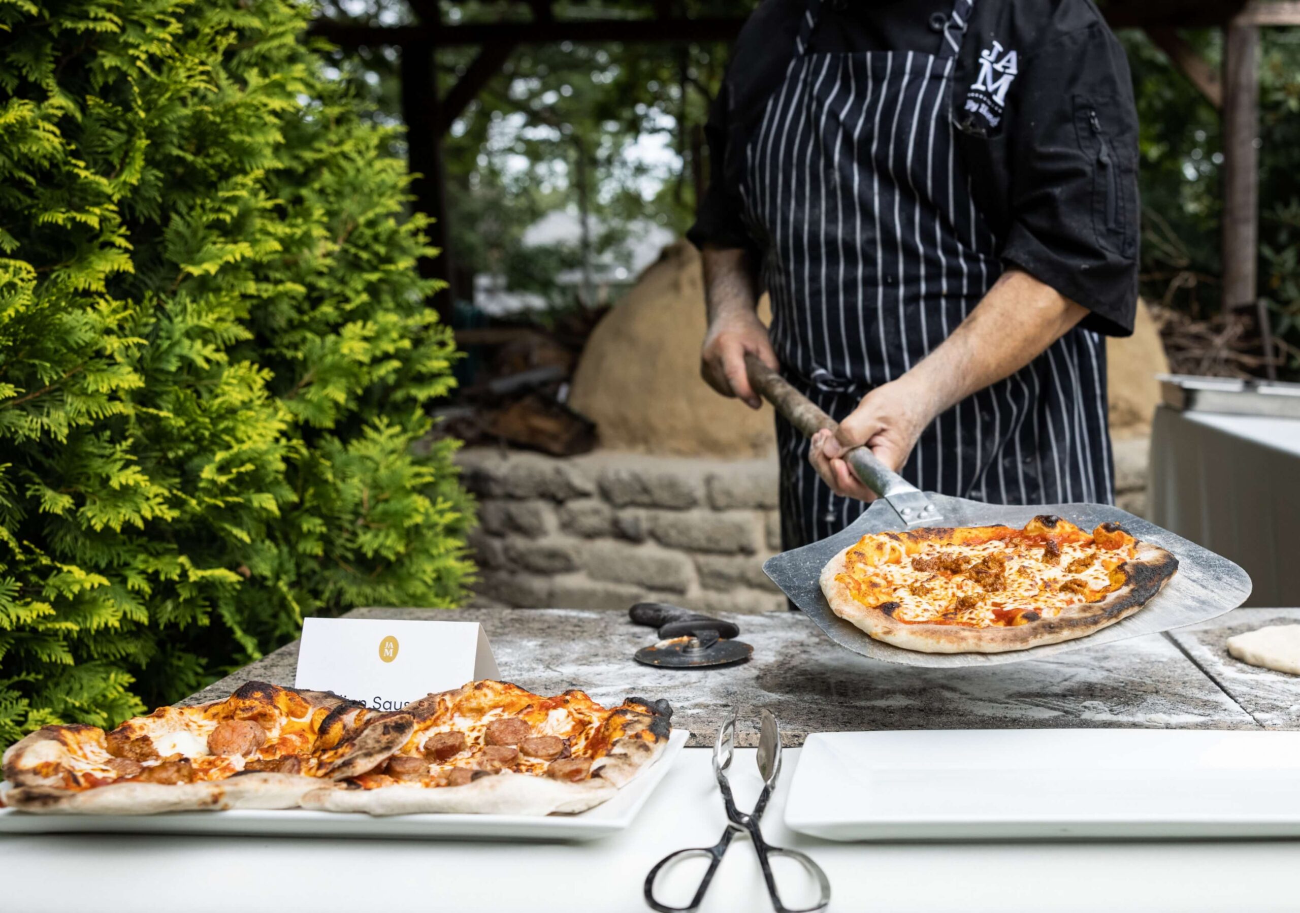 Person in a striped apron holding a pizza on a peel next to a brick oven outdoors. Nearby are tongs and additional pizzas on a table with a small card labeled 