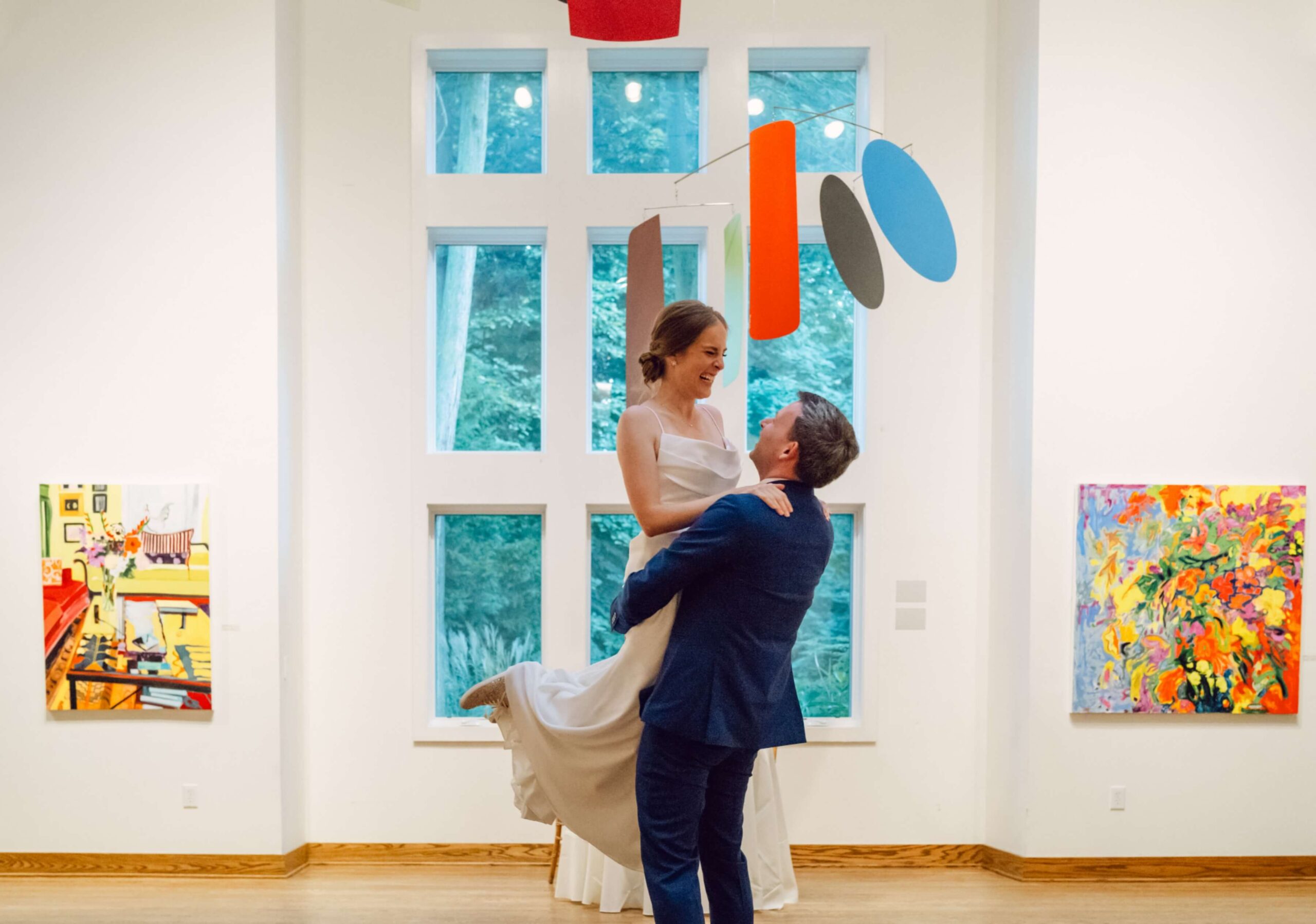 A groom lifts a bride in a modern art gallery, surrounded by colorful paintings and hanging sculptures.