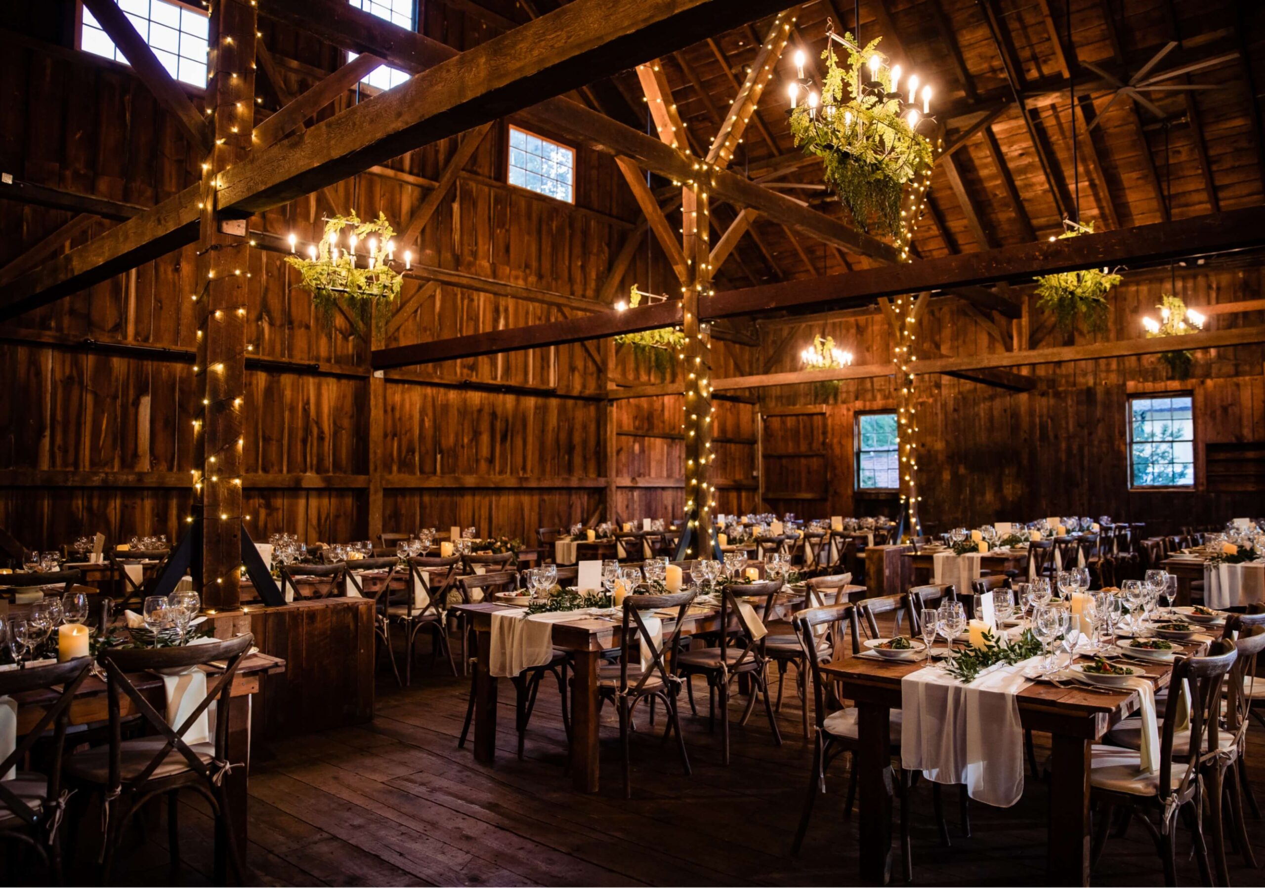 A rustic barn setting with long tables arranged for a formal event. Tables are decorated with white cloths, greenery, and candles. Warm string lights adorn the wooden beams above.
