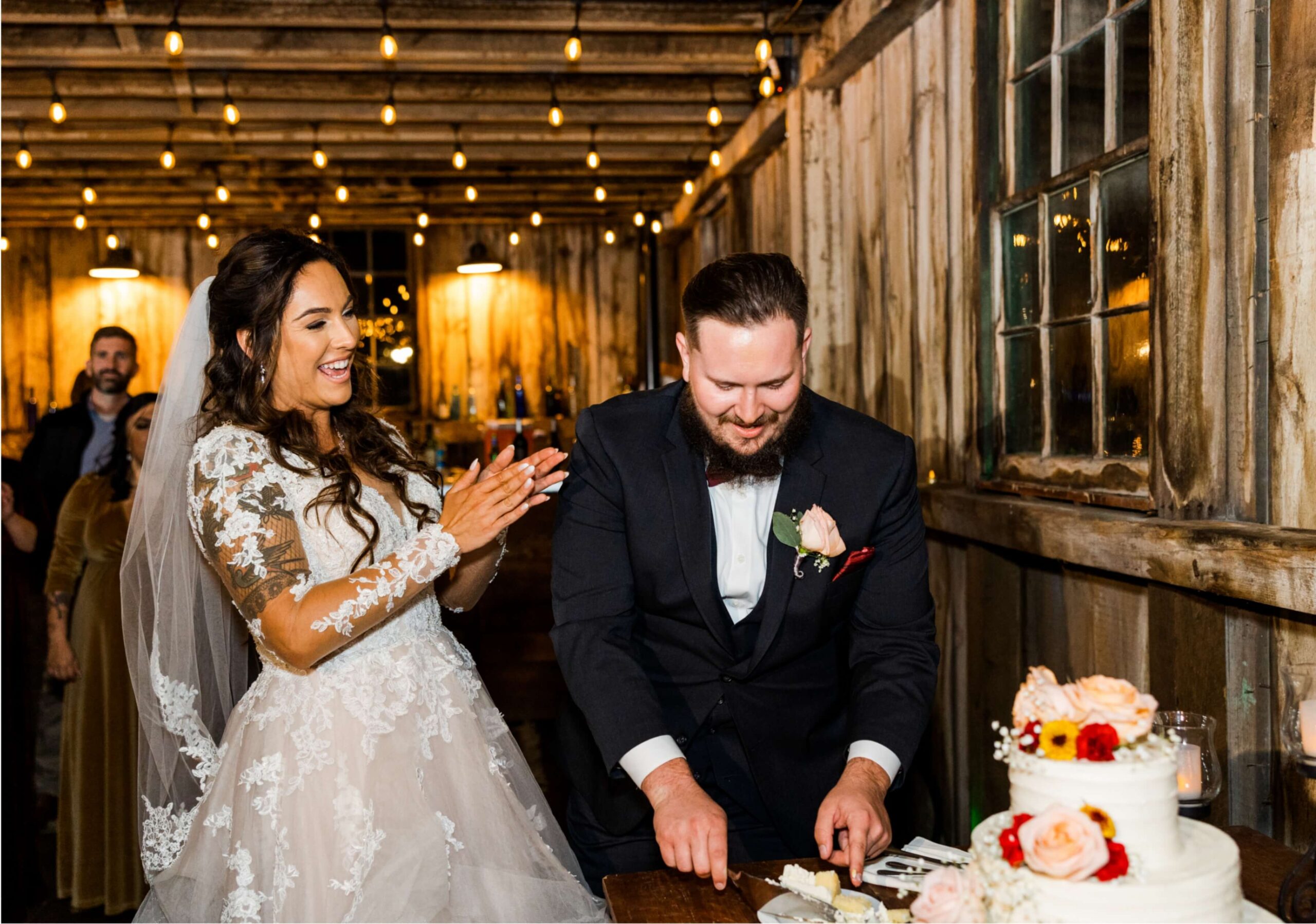 A bride and groom are cutting a wedding cake together. The bride is clapping and smiling. The setting is a rustic venue with string lights.