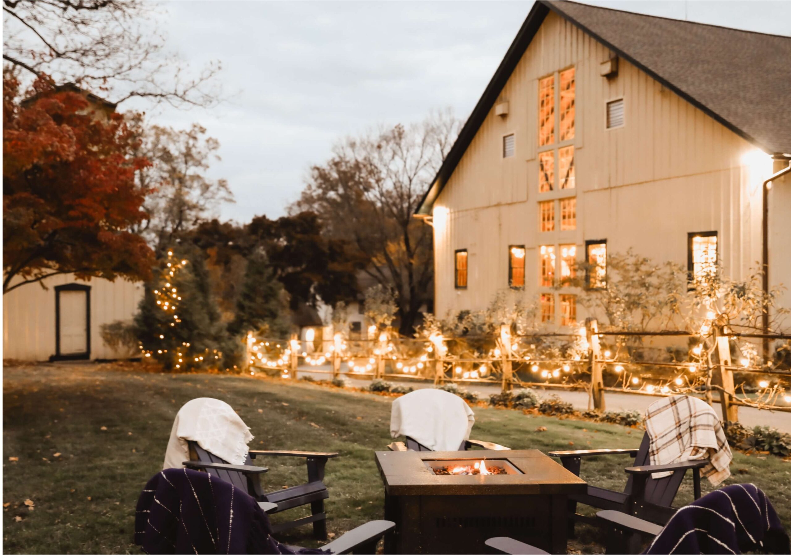 A cozy outdoor setting with Adirondack chairs around a fire pit, a barn decorated with string lights in the background, and trees with autumn foliage.