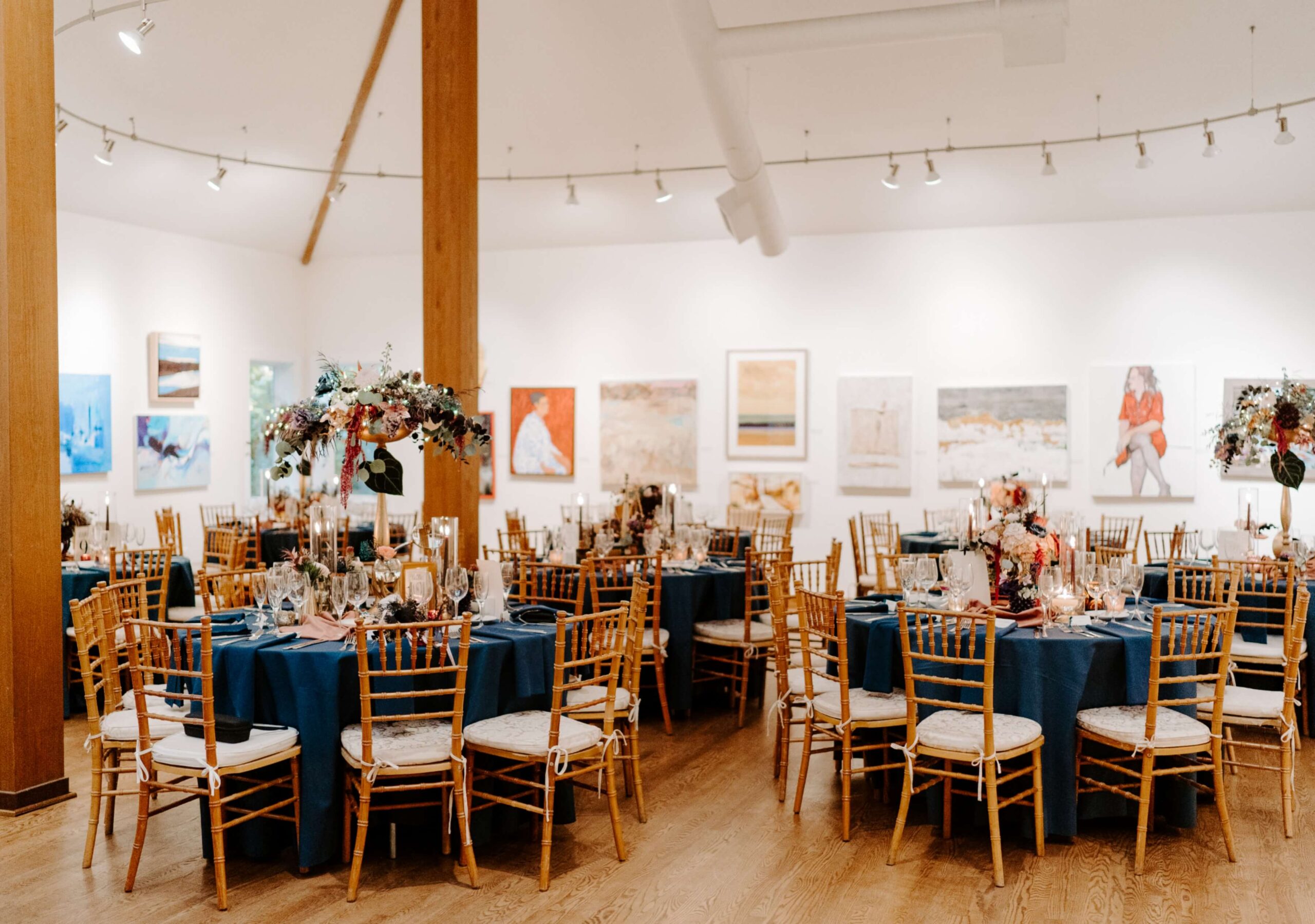 Elegant event setup with round tables draped in blue tablecloths, adorned with floral centerpieces, surrounded by wooden chairs. Artworks hang on white walls in a well-lit room.