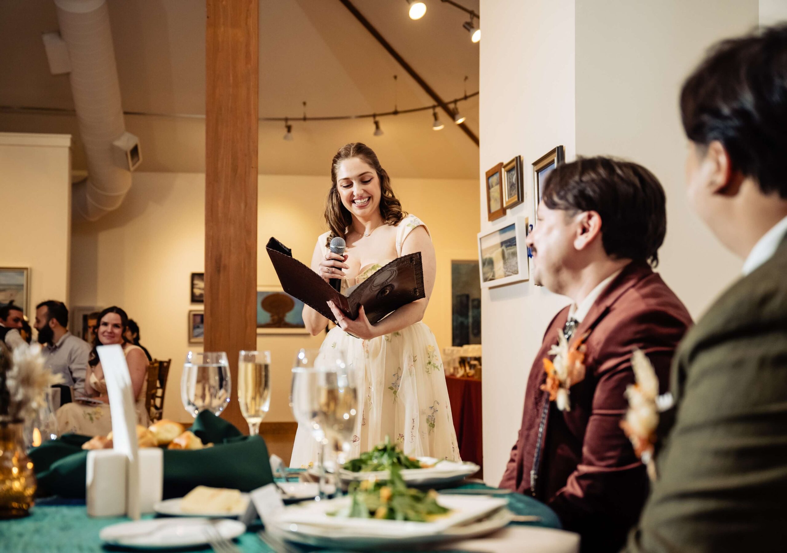 A woman in a dress speaks into a microphone, holding a folder, while two seated individuals listen at a dining event.