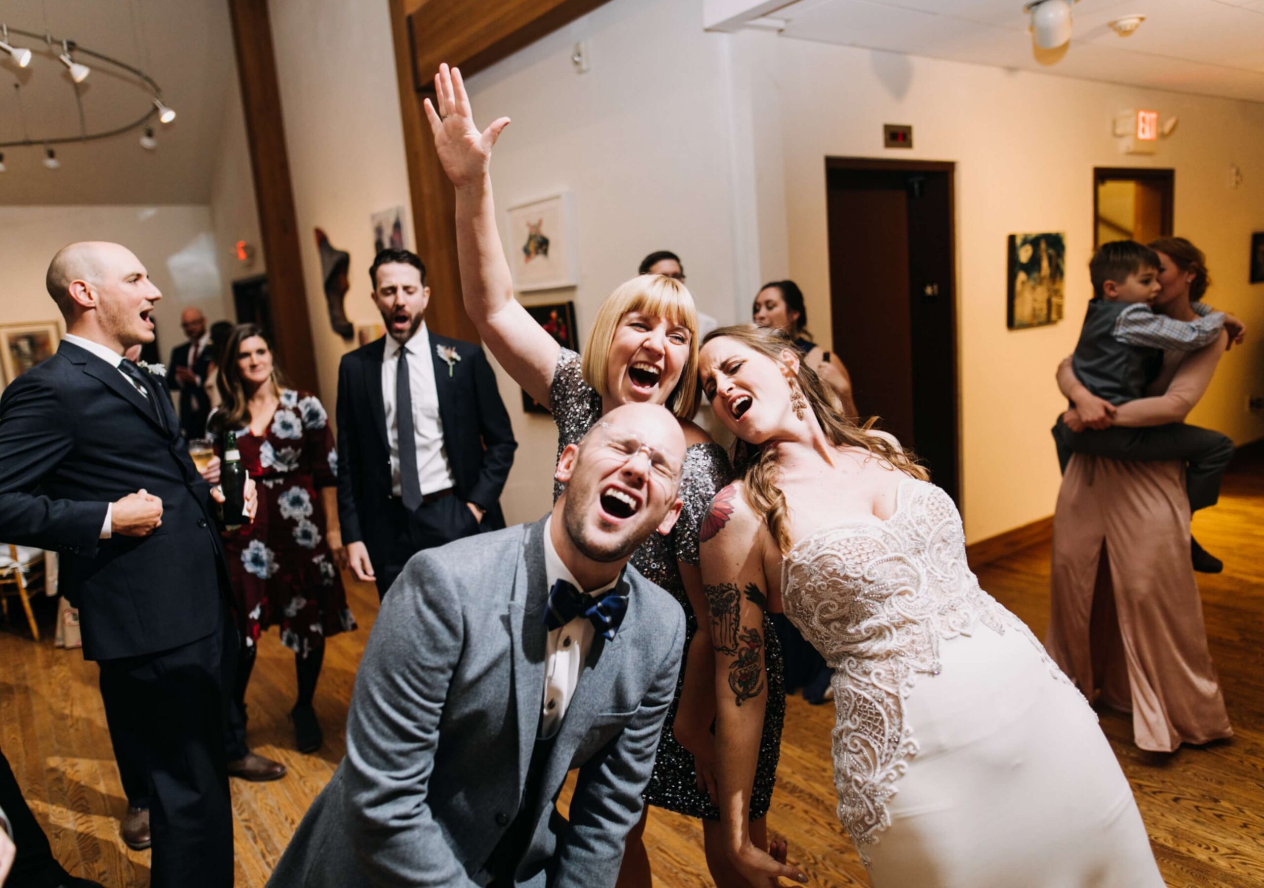 People dancing and laughing at a lively indoor event; a groom and bride stand in the foreground, joyfully leaning forward.