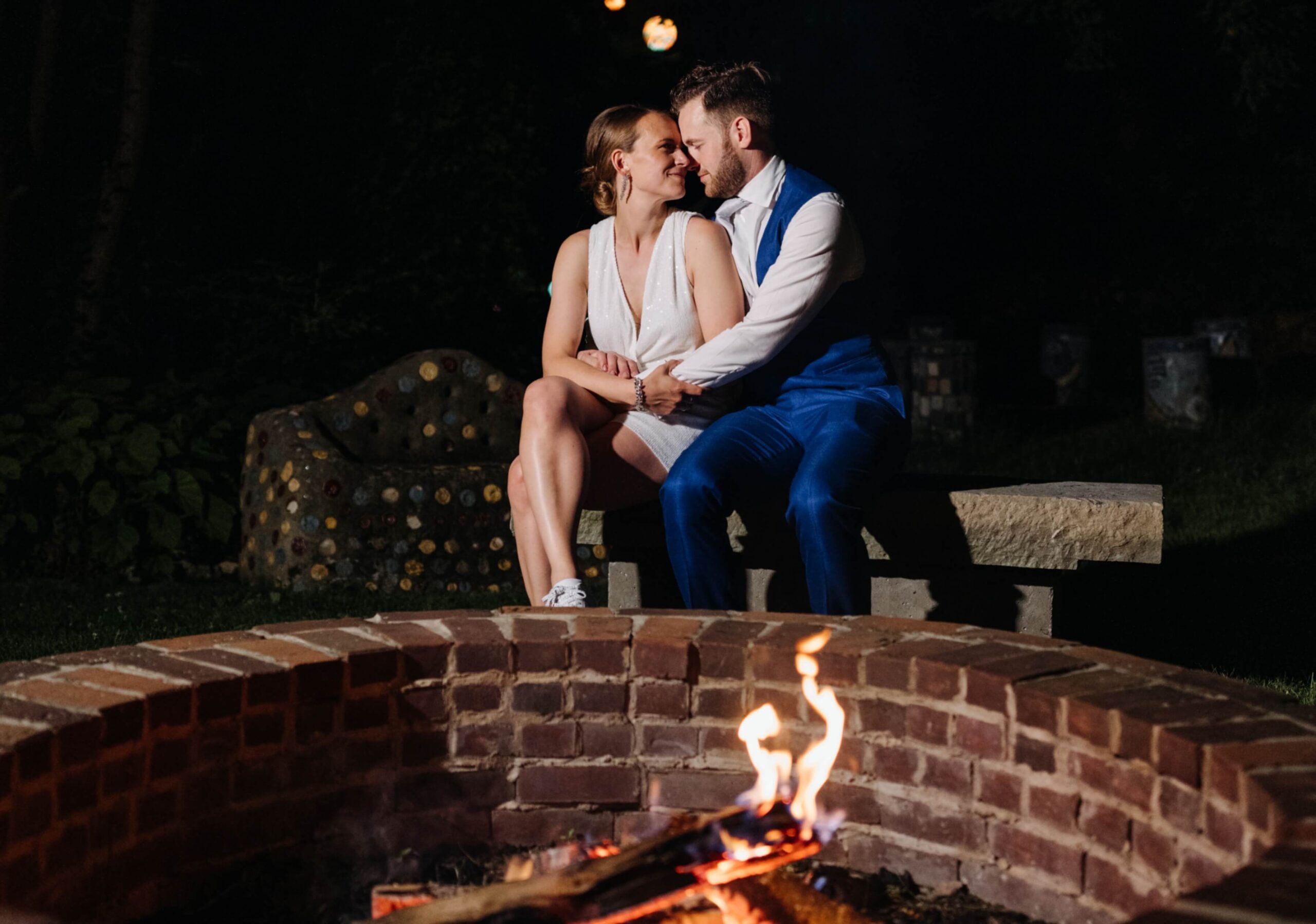 A couple sits on a stone bench by a fire pit at night. The woman wears a white dress, and the man wears a blue suit. They are looking at each other affectionately.