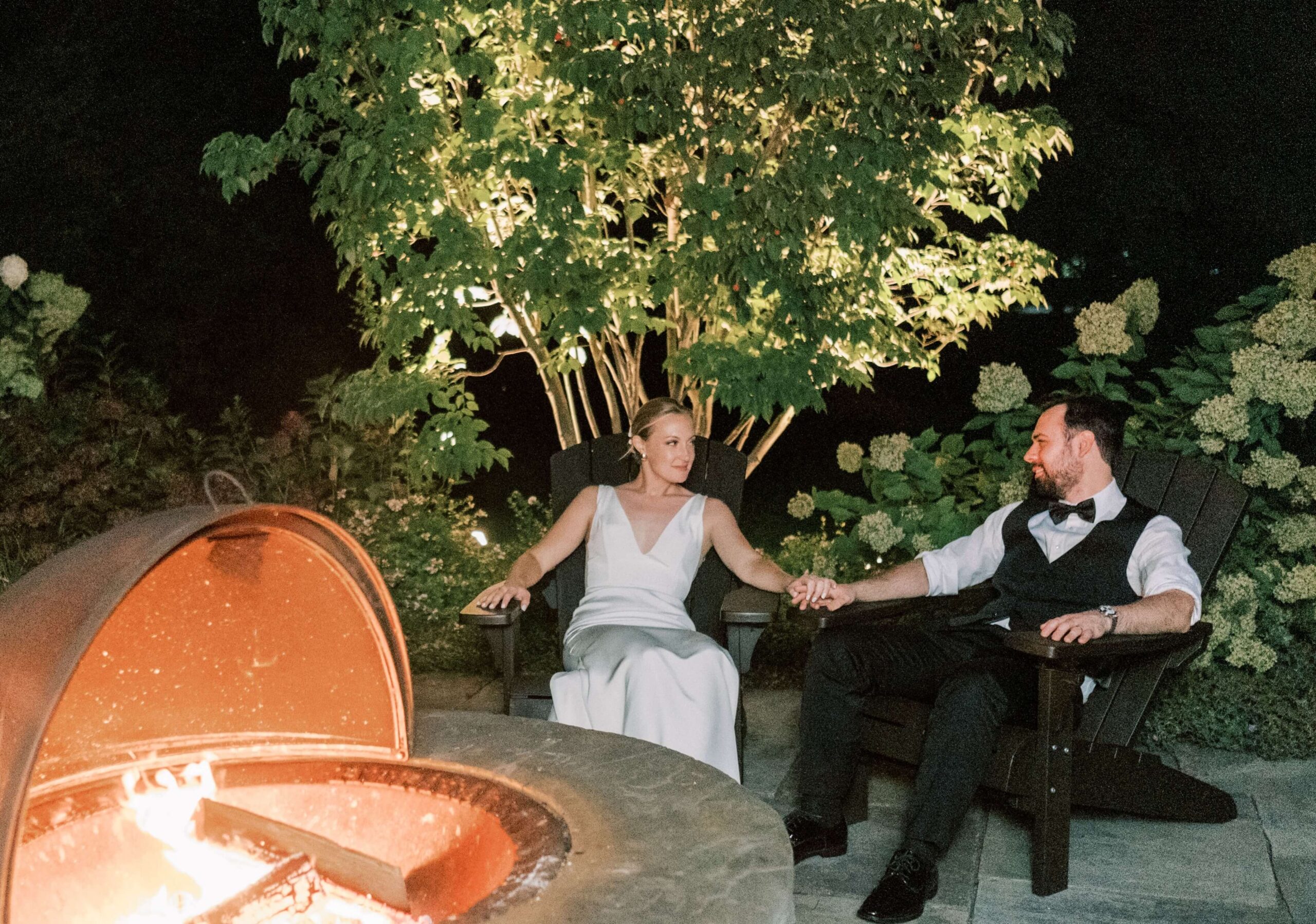 A couple dressed in formal attire sits holding hands in front of a fire pit surrounded by lush greenery at night.