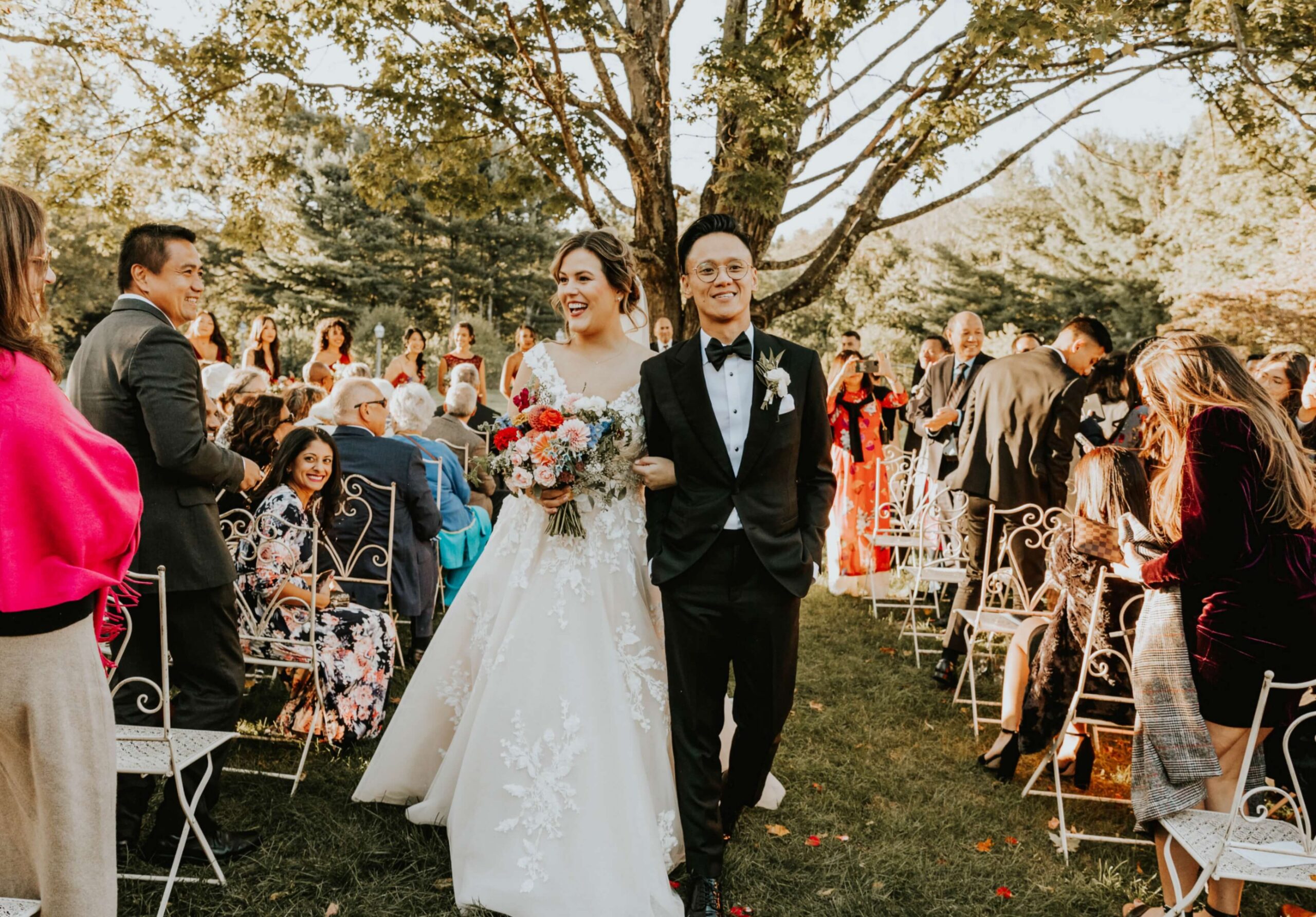 A bride and groom walk down an outdoor aisle, smiling, surrounded by seated guests. The bride holds a bouquet, and tree branches are visible in the background.
