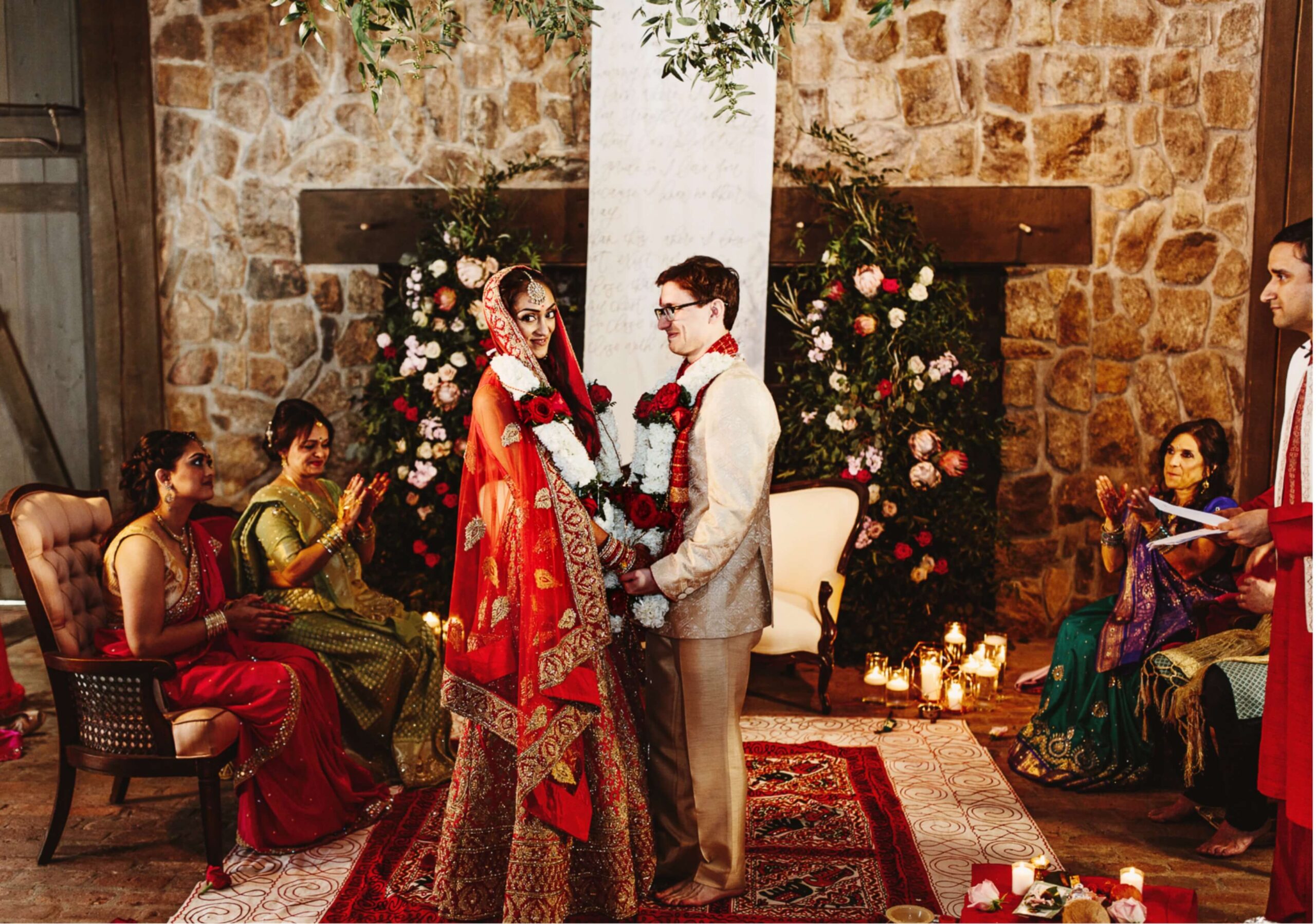 A couple in wedding attire stands facing each other during a ceremony. They are surrounded by floral decorations and seated guests, with a stone wall and candles in the background.