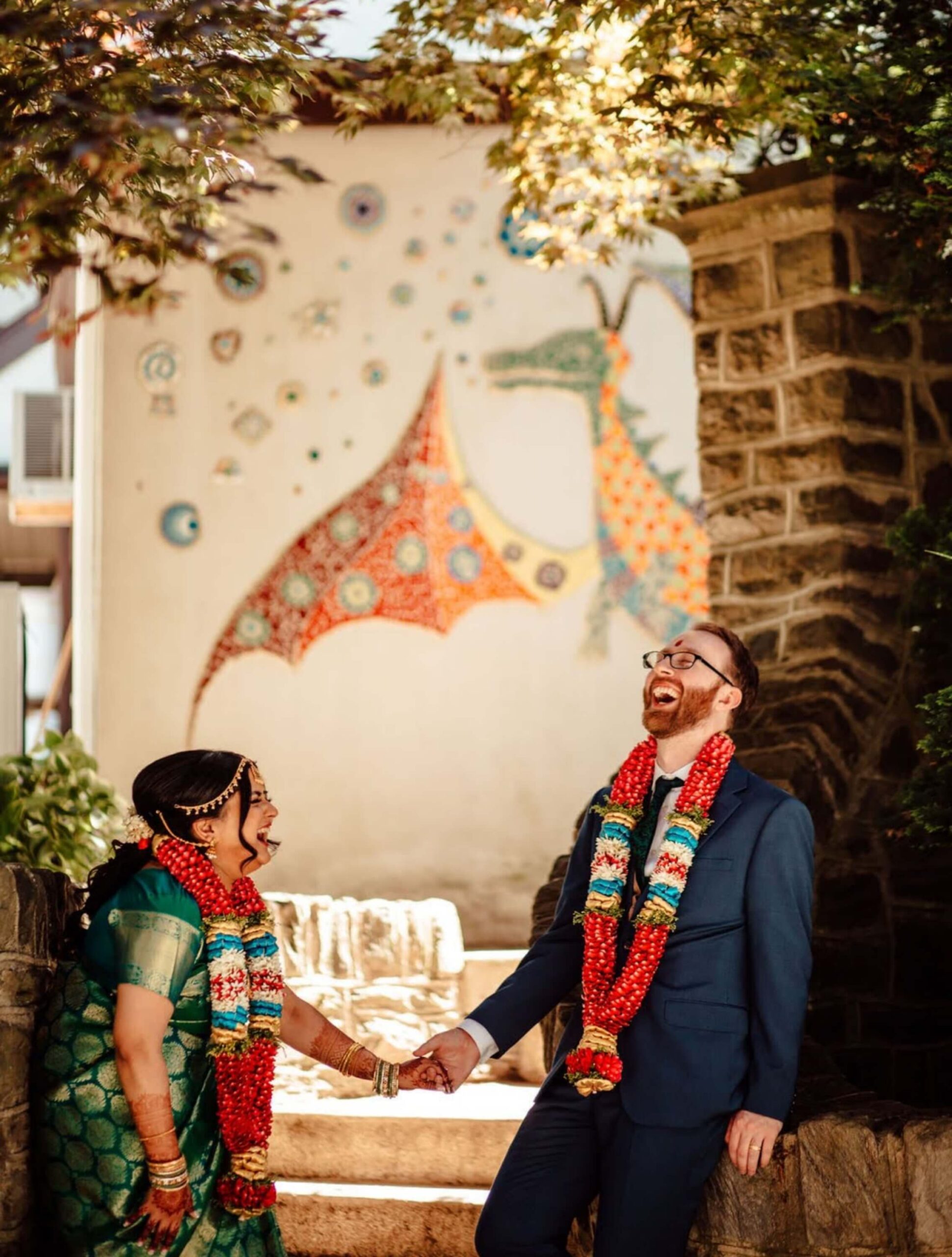 A couple, wearing colorful traditional attire and garlands, laughing while holding hands. They stand near a stone wall with a vibrant dragon mural in the background.