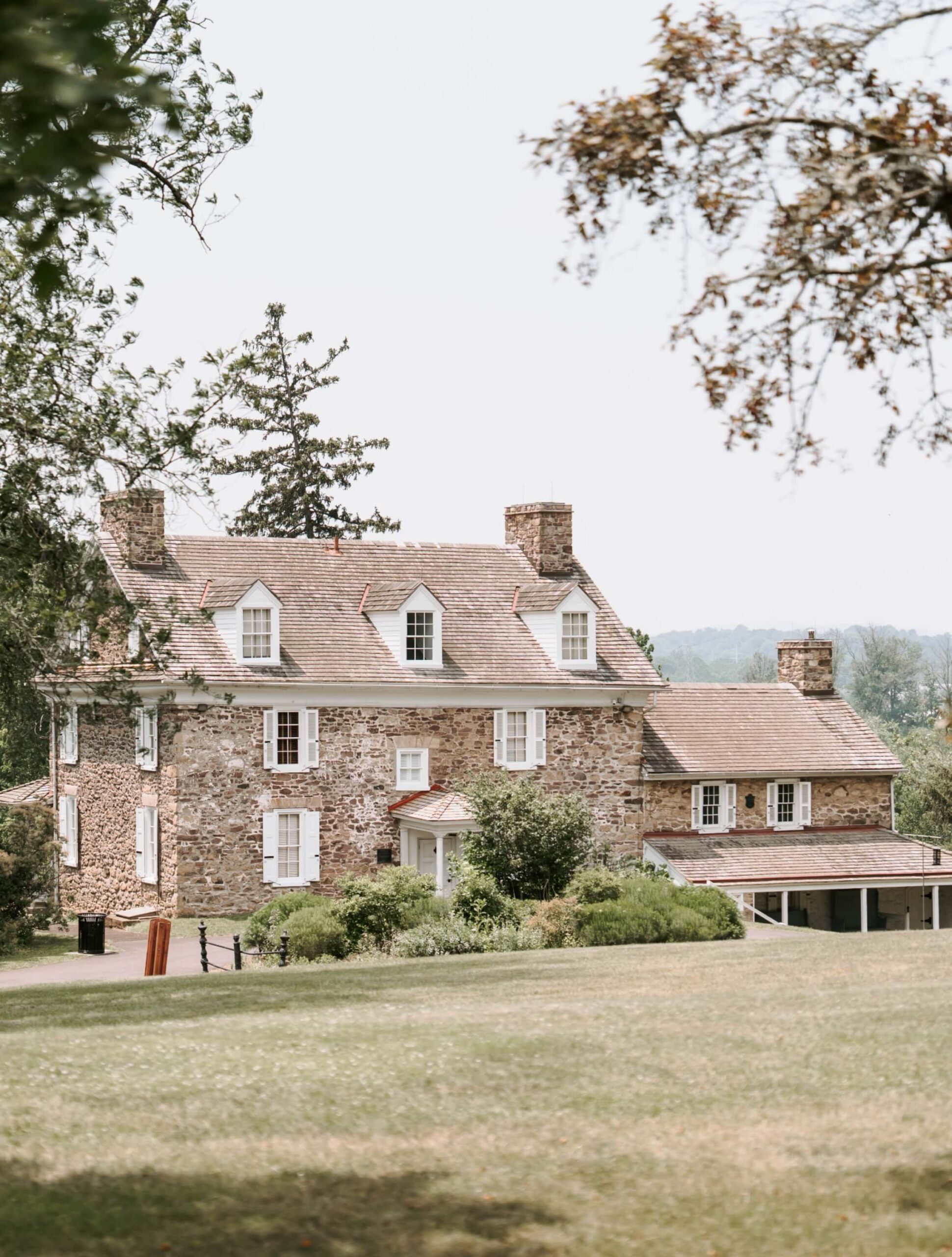 A large stone house with multiple chimneys and dormer windows sits amidst a green lawn and trees under a clear sky.
