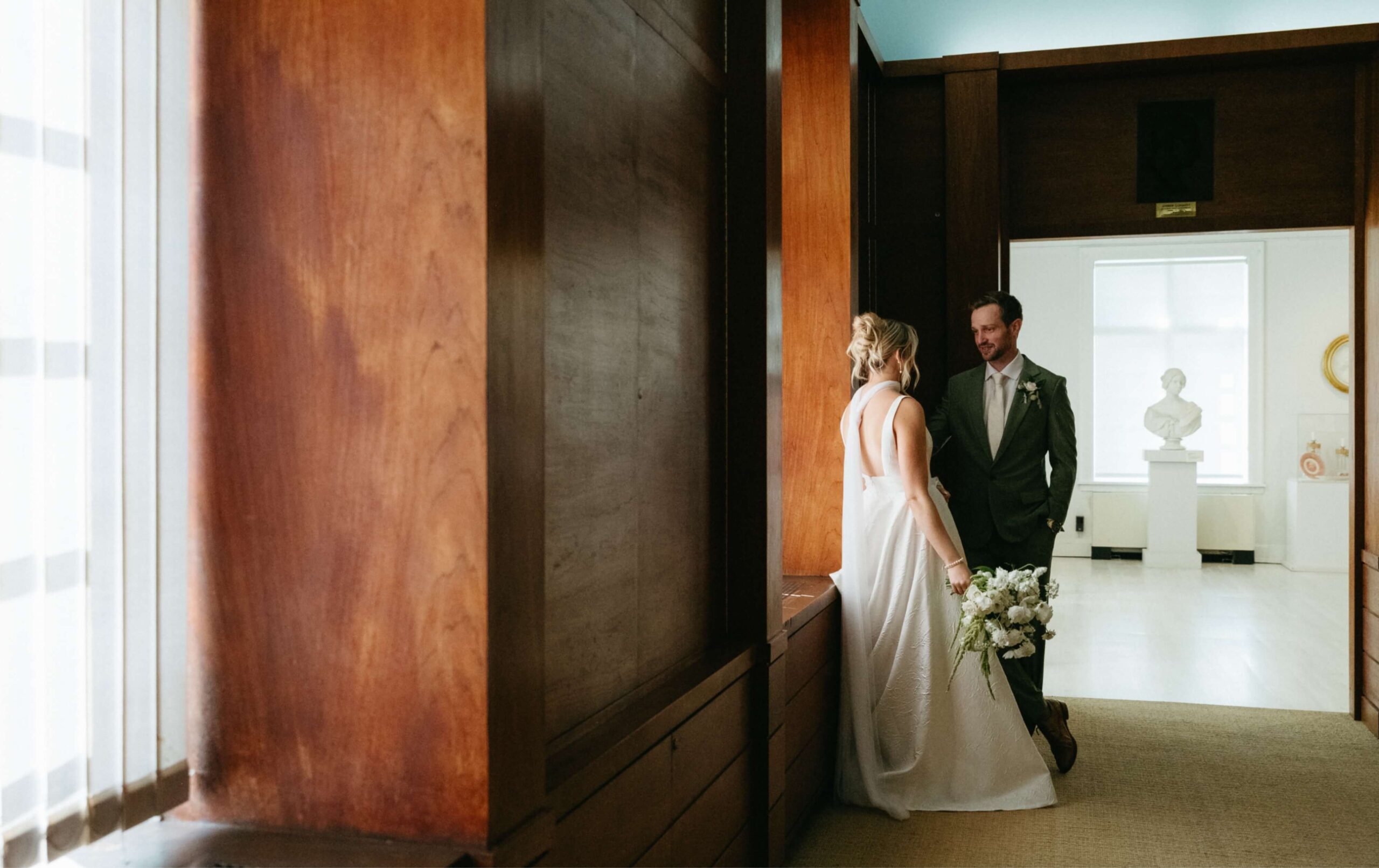 Bride in a white dress holds a bouquet while standing in a hallway with a groom in a suit. They are near a large wooden panel and a window.
