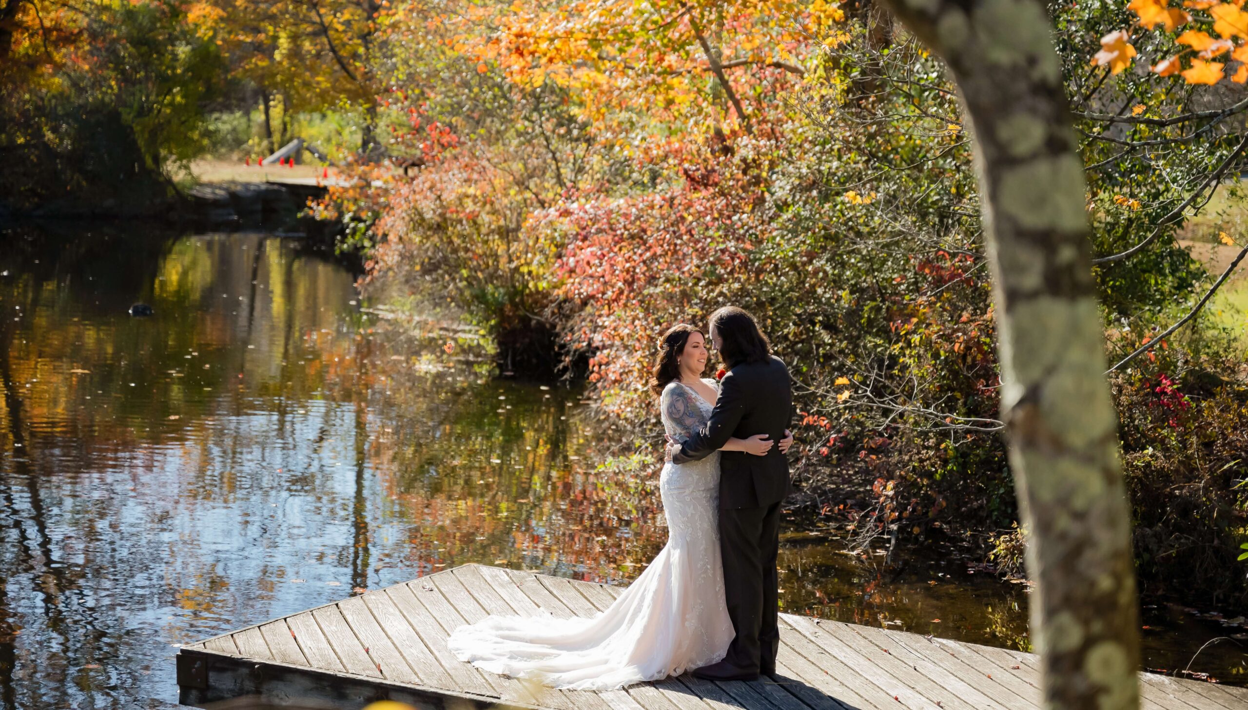 A couple in wedding attire stands on a wooden dock surrounded by autumnal trees, near a calm reflective pond.
