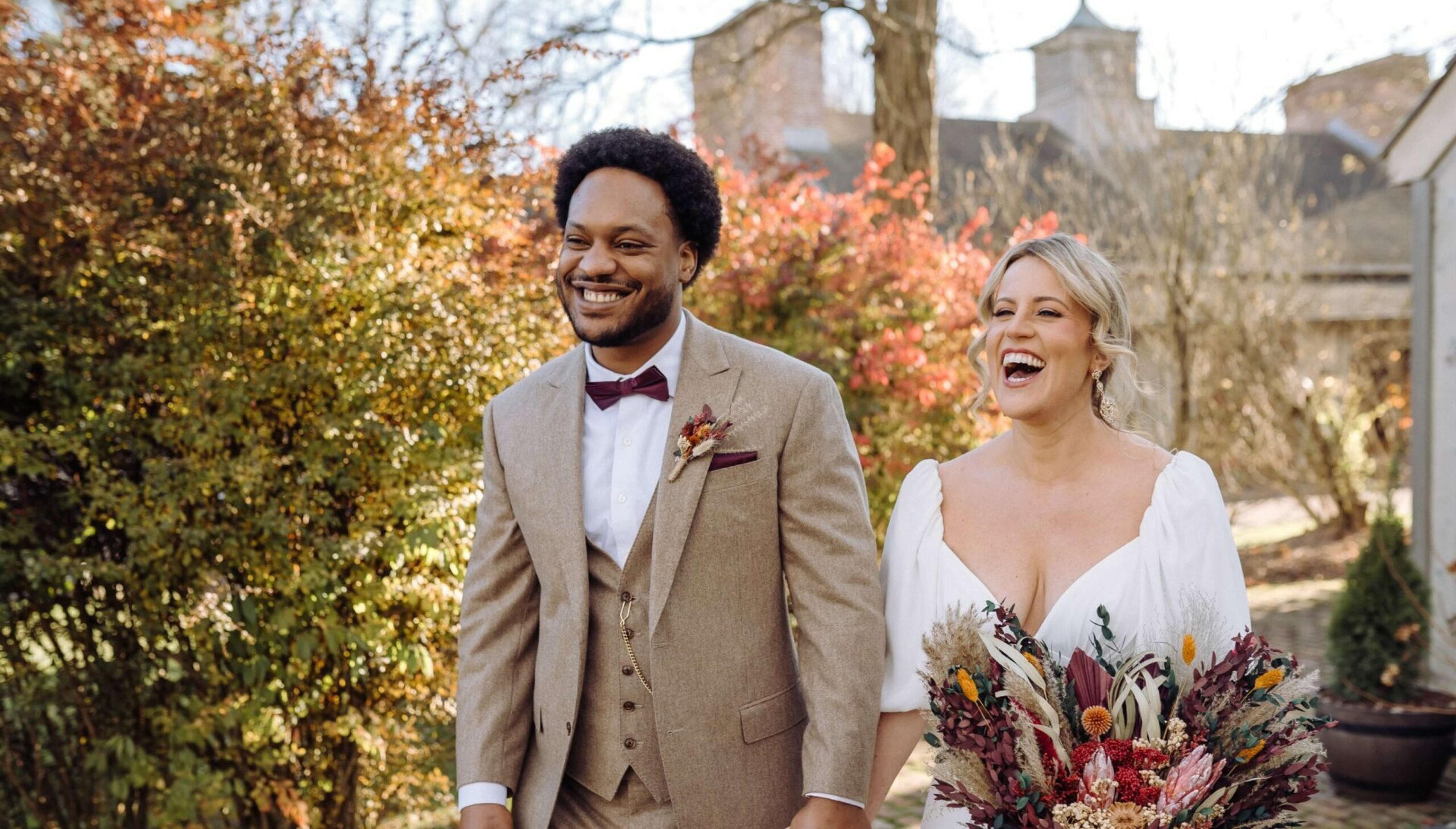 A couple smiles while walking outdoors. The man wears a tan suit and red bow tie; the woman wears a white dress and carries a colorful bouquet. Autumn foliage is in the background.