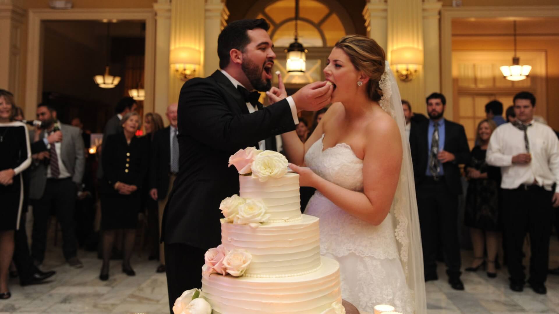 Bride and groom eating wedding cake together in a large hall, surrounded by guests.