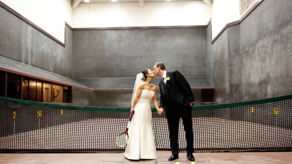 Bride and groom kissing on an indoor tennis court, standing in front of a net. Bride holds a tennis racket.