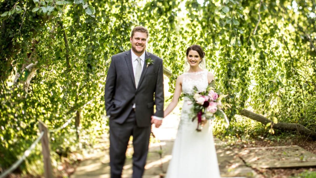 A bride and groom smiling and holding hands while walking on a wooden path surrounded by lush greenery.