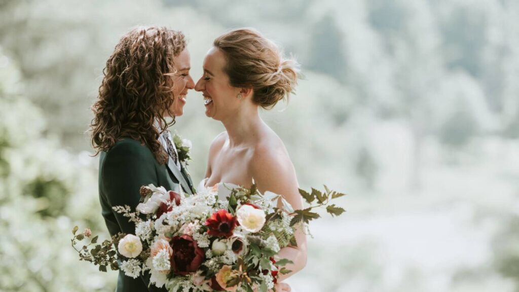 Two people in wedding attire smile and hold a floral bouquet, standing outdoors with a blurred green background.