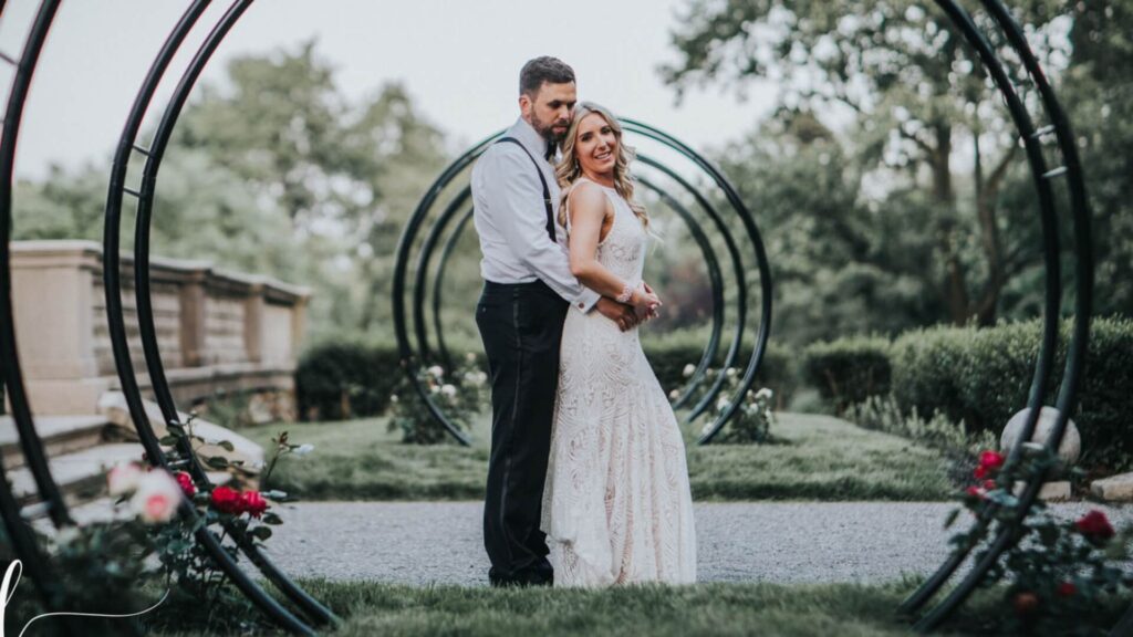 A couple stands embracing in a garden, surrounded by circular metal arches and greenery.