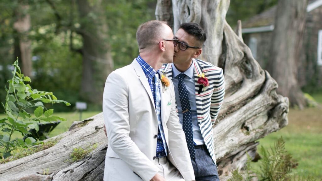 Two men in suits share a kiss while sitting and leaning against a large piece of driftwood in a garden setting.
