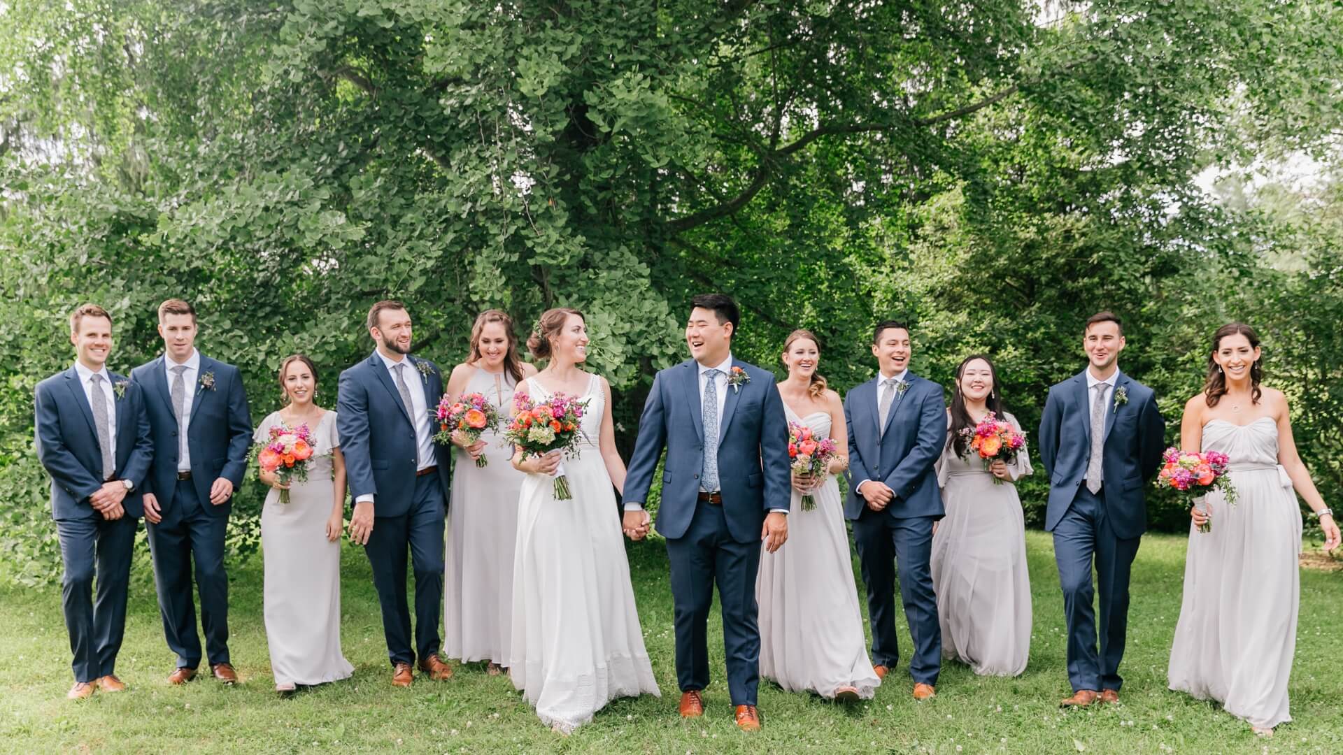 A wedding party stands outdoors, featuring the bride and groom in the center. Bridesmaids and groomsmen in coordinated outfits hold bouquets, with a backdrop of green trees and grass.
