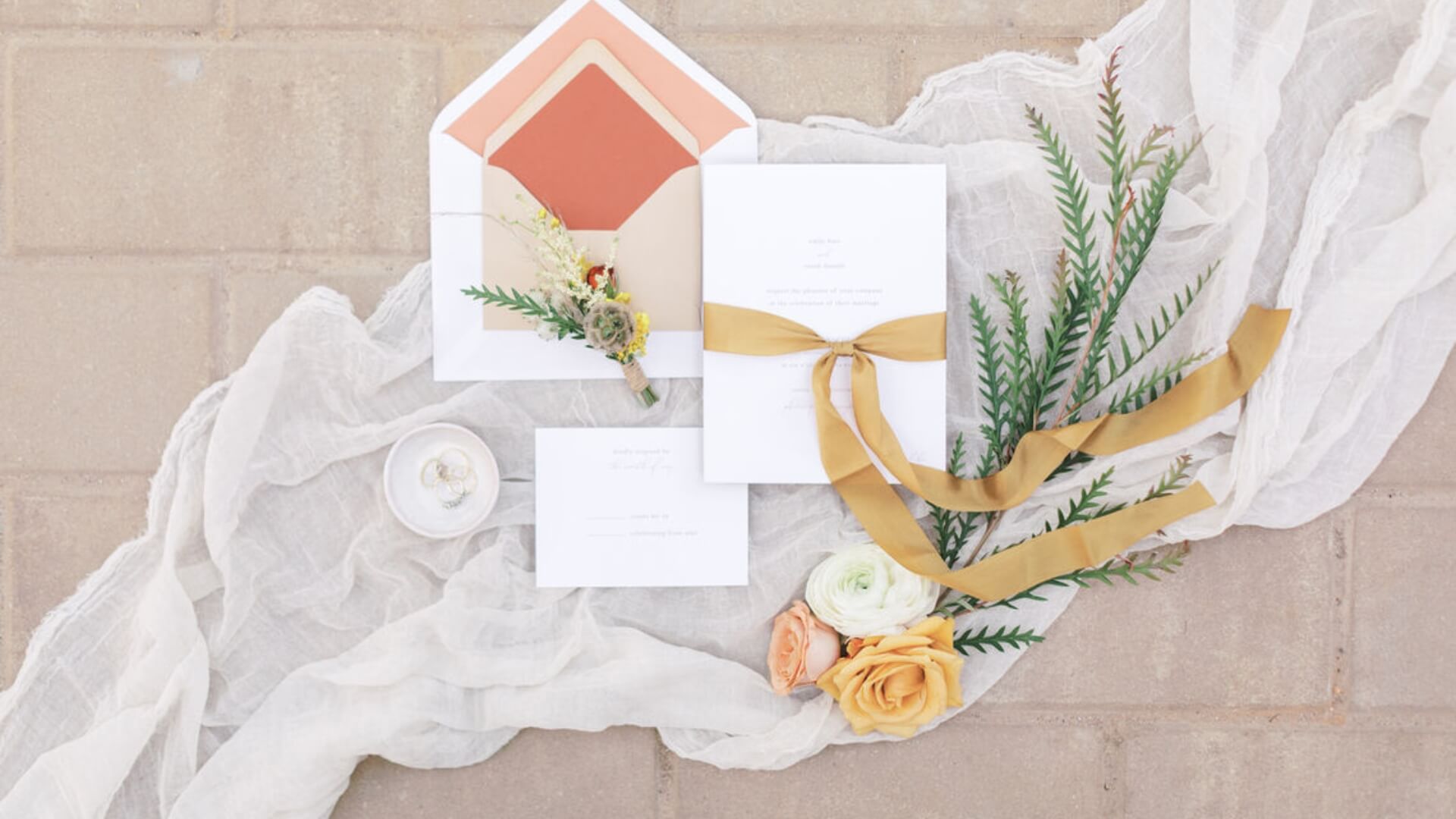 Wedding invitation set with orange, beige, and white envelopes, flowers, and a gold ribbon on a textured background with sheer fabric.