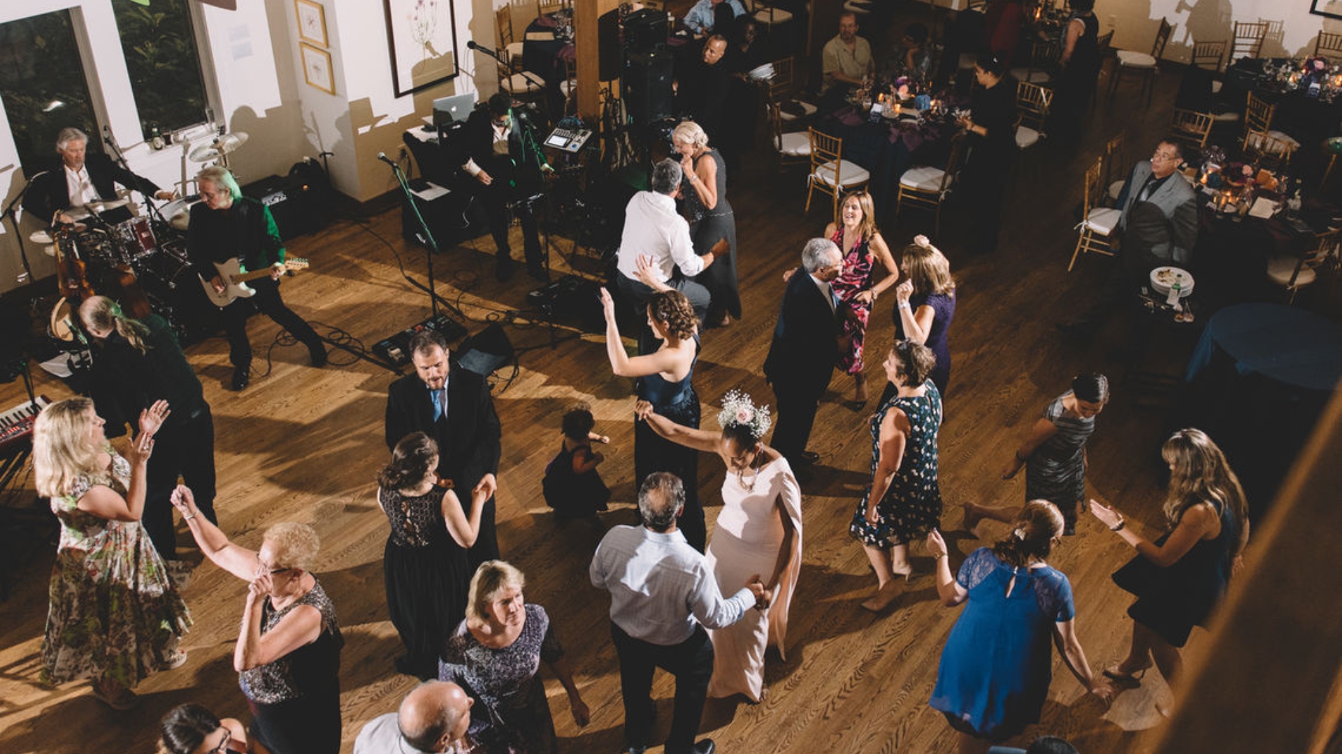 Guests dance at a formal event with live music in a spacious room with wooden flooring and decorated tables.