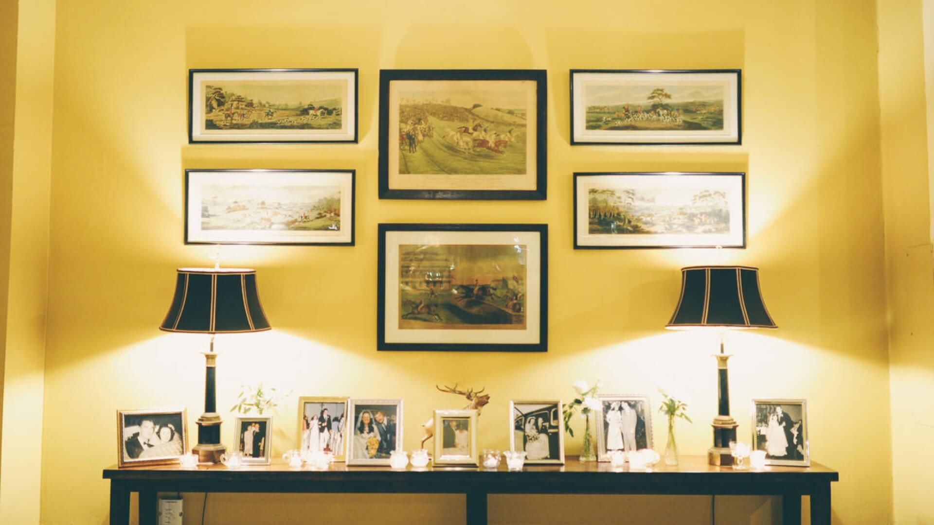 A yellow wall with framed landscape paintings above a console table. The table has two lamps and several framed photographs.