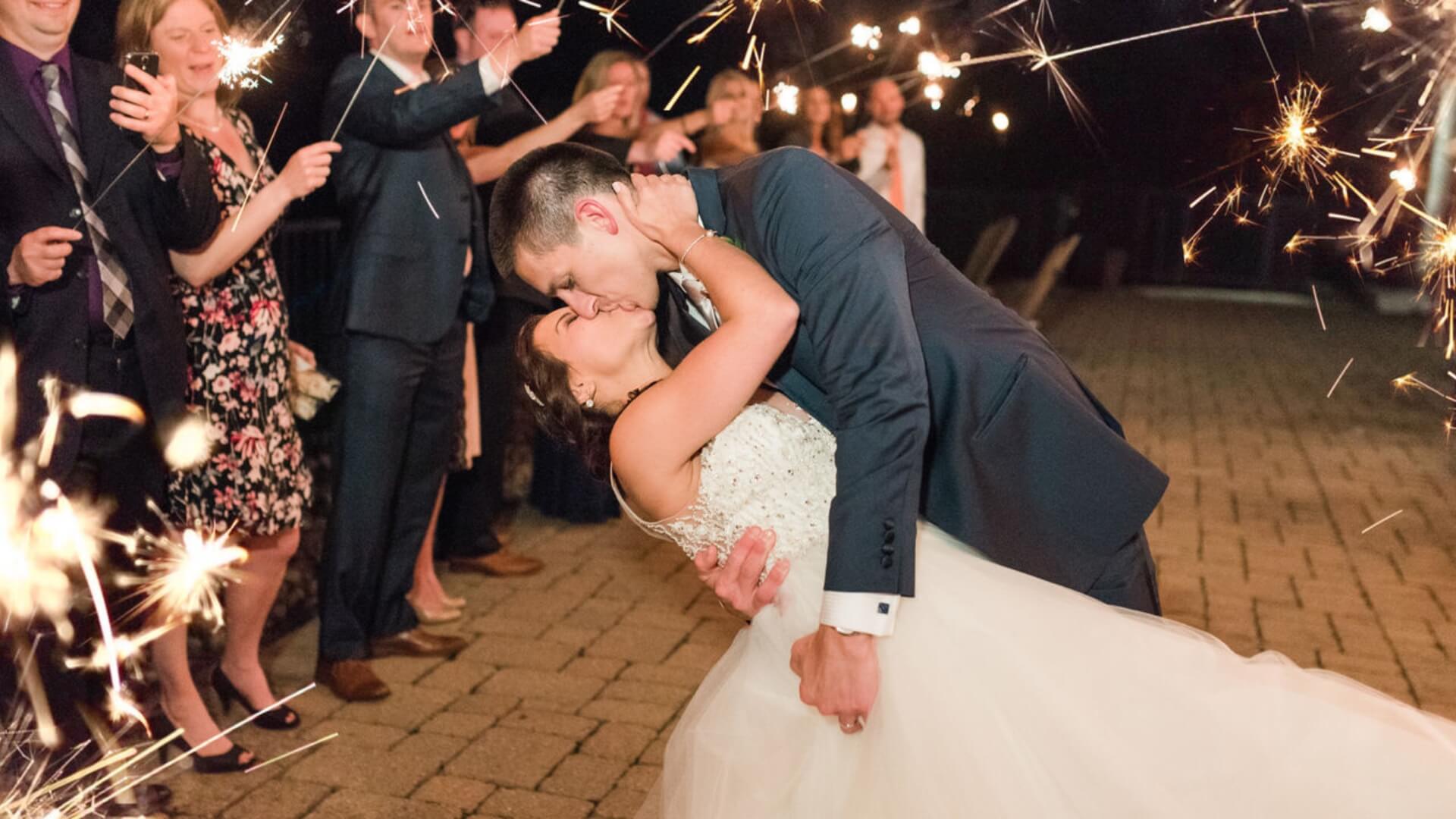 A newlywed couple shares a kiss in a dip pose amidst a celebratory crowd holding sparklers at night.