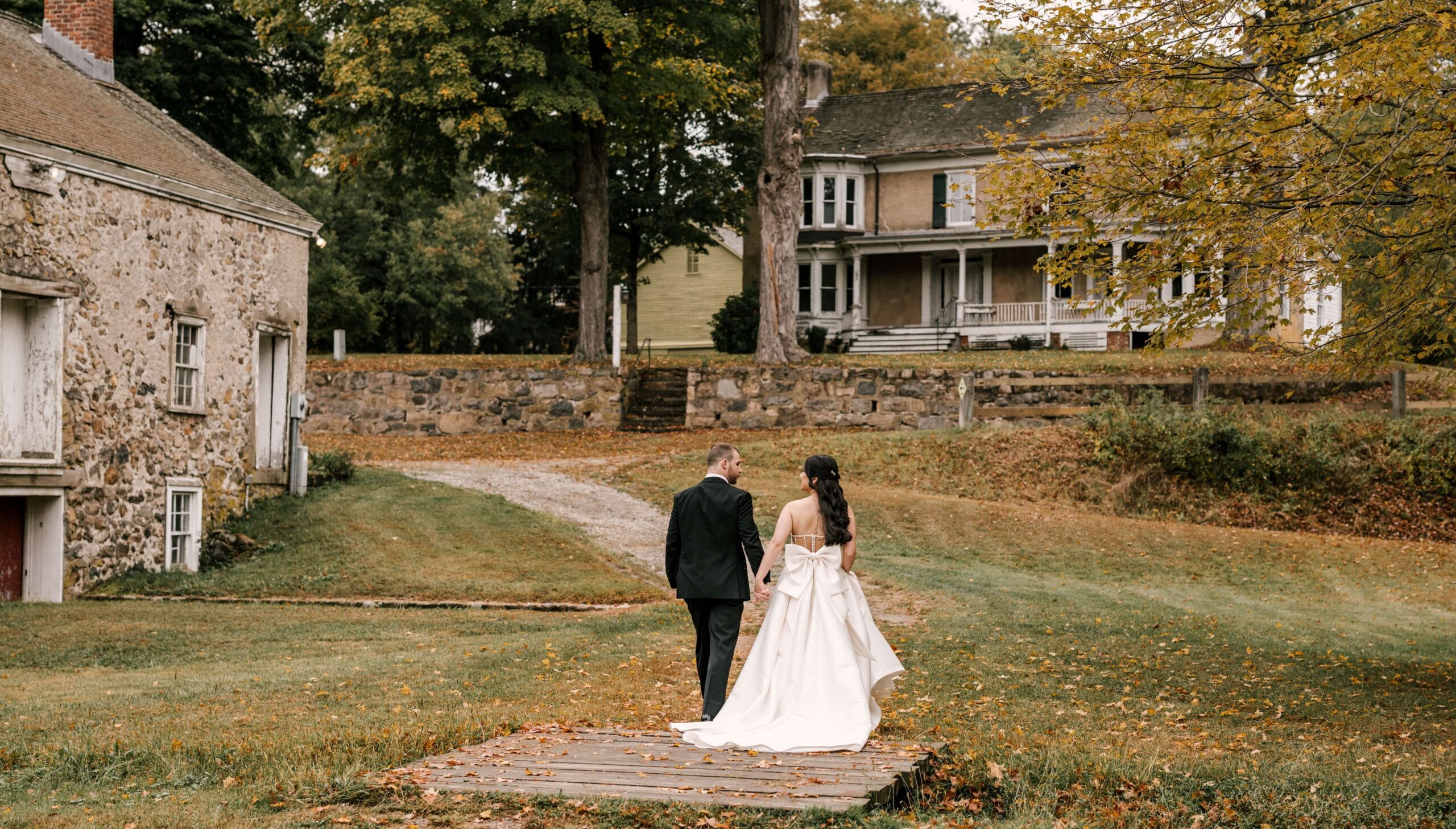 A couple in formal attire walks hand in hand on a wooden path in a rustic setting with grass, trees, and two stone houses.