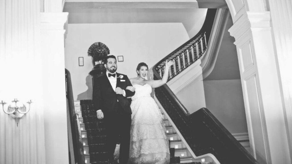 Bride and groom in formal attire descend a staircase, smiling and waving.
