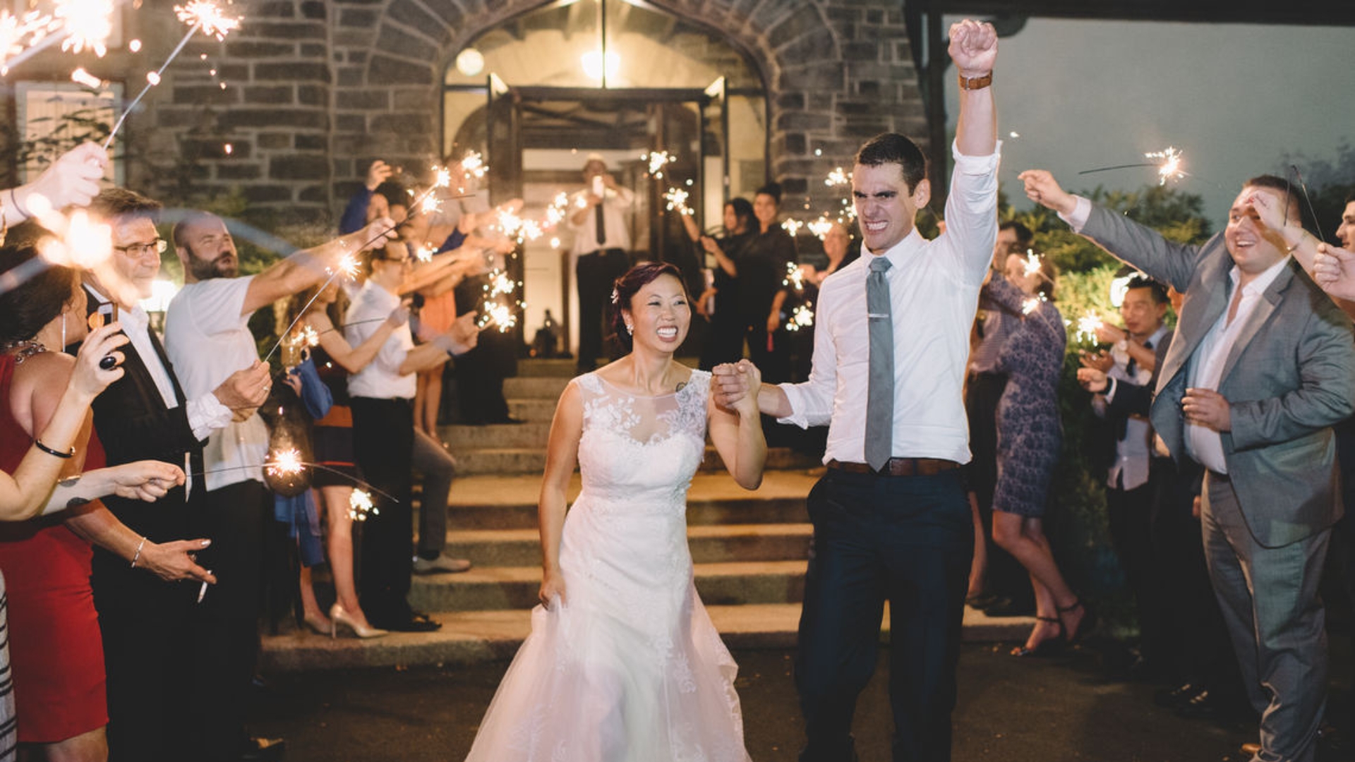 A bride and groom hold hands and celebrate as they walk through a group of people holding sparklers outside a building.