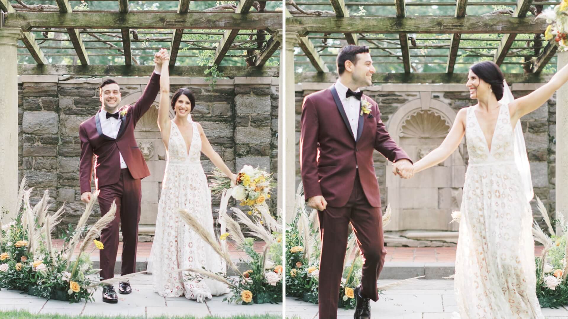 A couple in wedding attire holds hands and celebrates under a wooden pergola. The groom wears a burgundy suit, and the bride is in a white dress holding a bouquet.