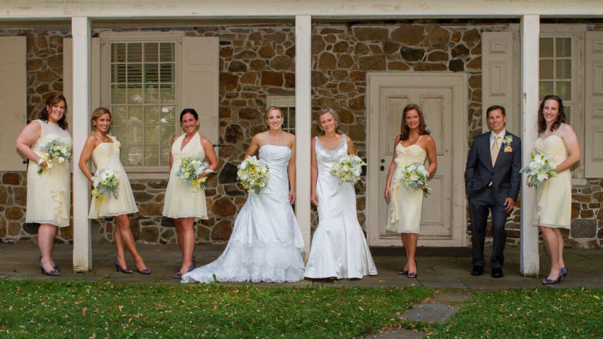 Eight people, two in wedding dresses, stand in front of a stone building. Three carry bouquets, and one wears a suit. They are outdoors on a grassy area.