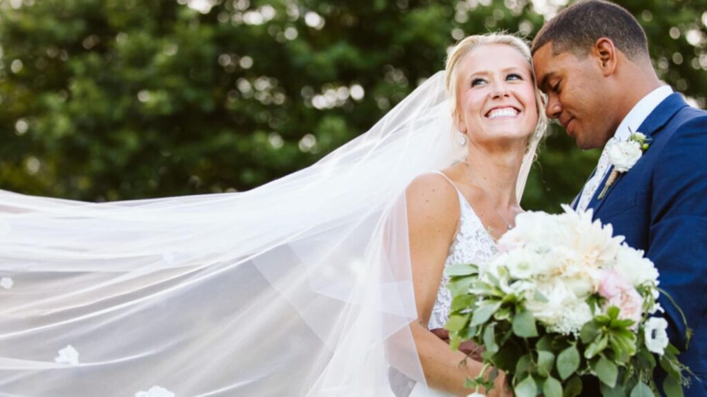 A bride and groom stand closely together outdoors. The bride smiles while holding a bouquet; her veil flows in the breeze. The groom leans his head against hers. Lush greenery is in the background.