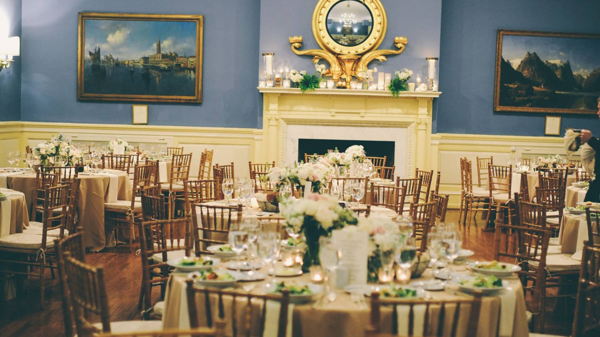 Elegant dining room with round tables set for an event, featuring floral centerpieces, wooden chairs, and a fireplace with a large mirror.