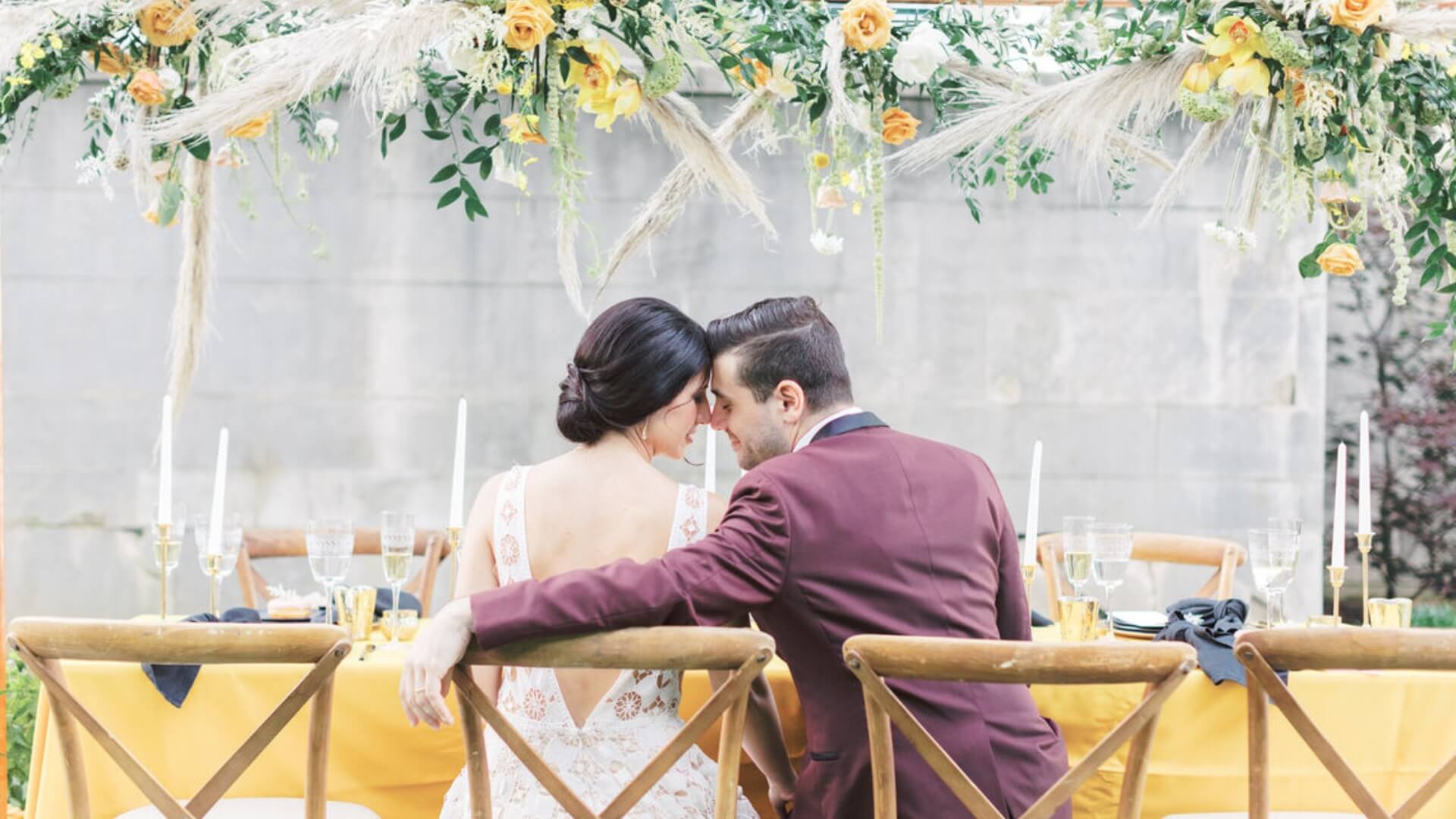 A couple sits closely at a decorated outdoor table adorned with candles, flowers, and greenery.