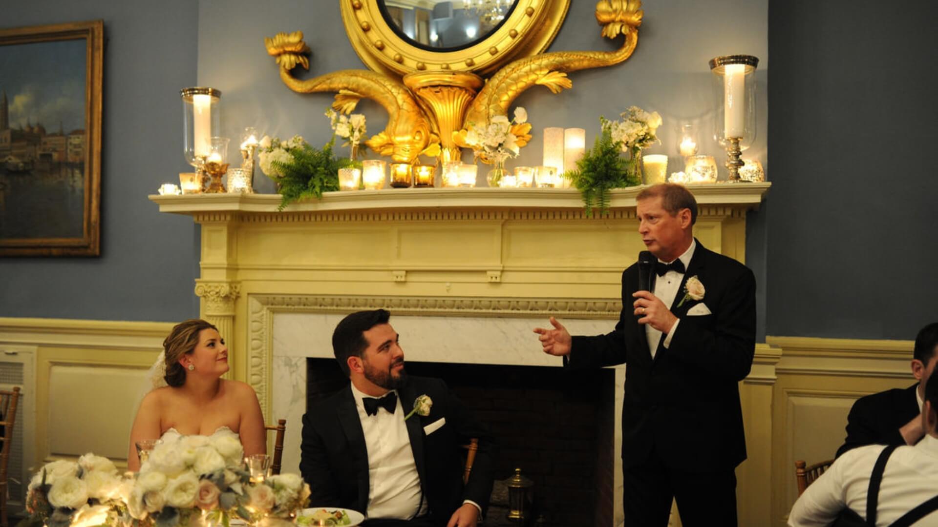 A man in a black tuxedo gives a speech with a microphone at a wedding reception, while the bride and groom, seated at the table, listen attentively. Ornate decorations adorn the mantel behind him.