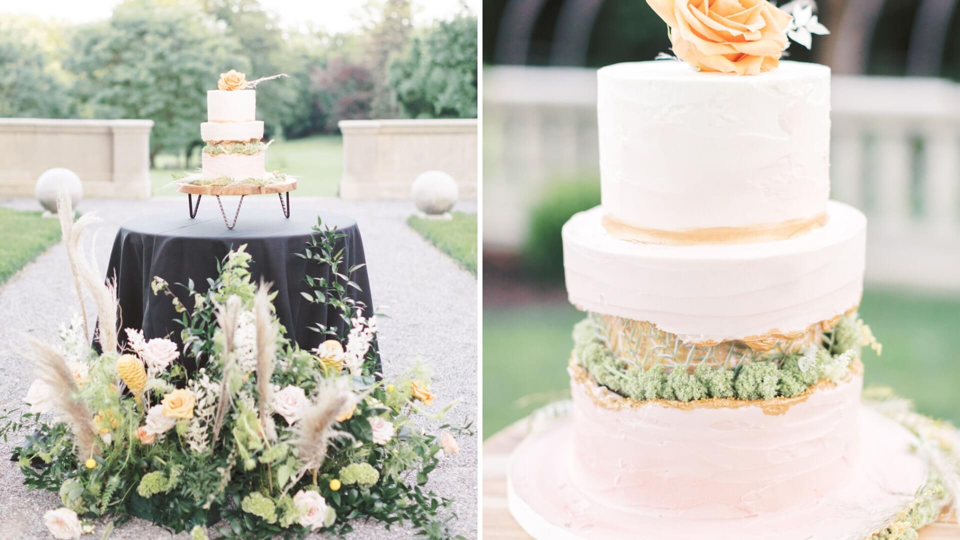 Two-tier white cake with orange flower on top, displayed on a round black table. The table is surrounded by greenery and peach flowers. The setting is outdoors with greenery in the background.