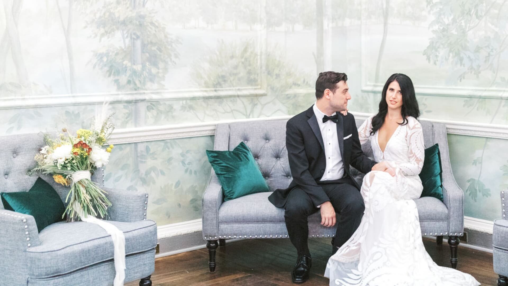 A couple in wedding attire sits on a gray sofa in a room with a nature-themed mural. A bouquet rests on another sofa next to them.