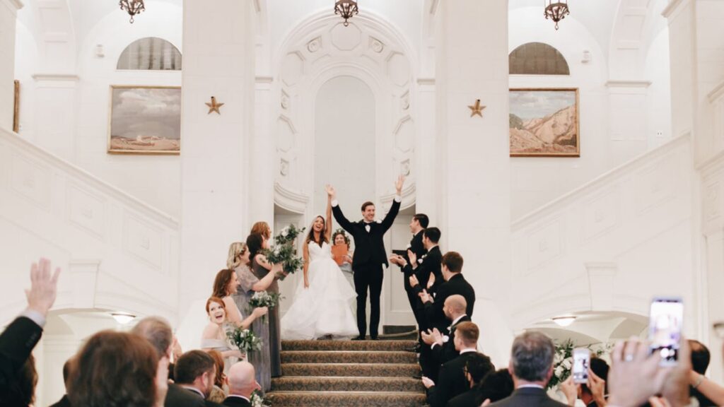 A newlywed couple holds hands, celebrating at the top of a staircase surrounded by cheering guests in formal attire.