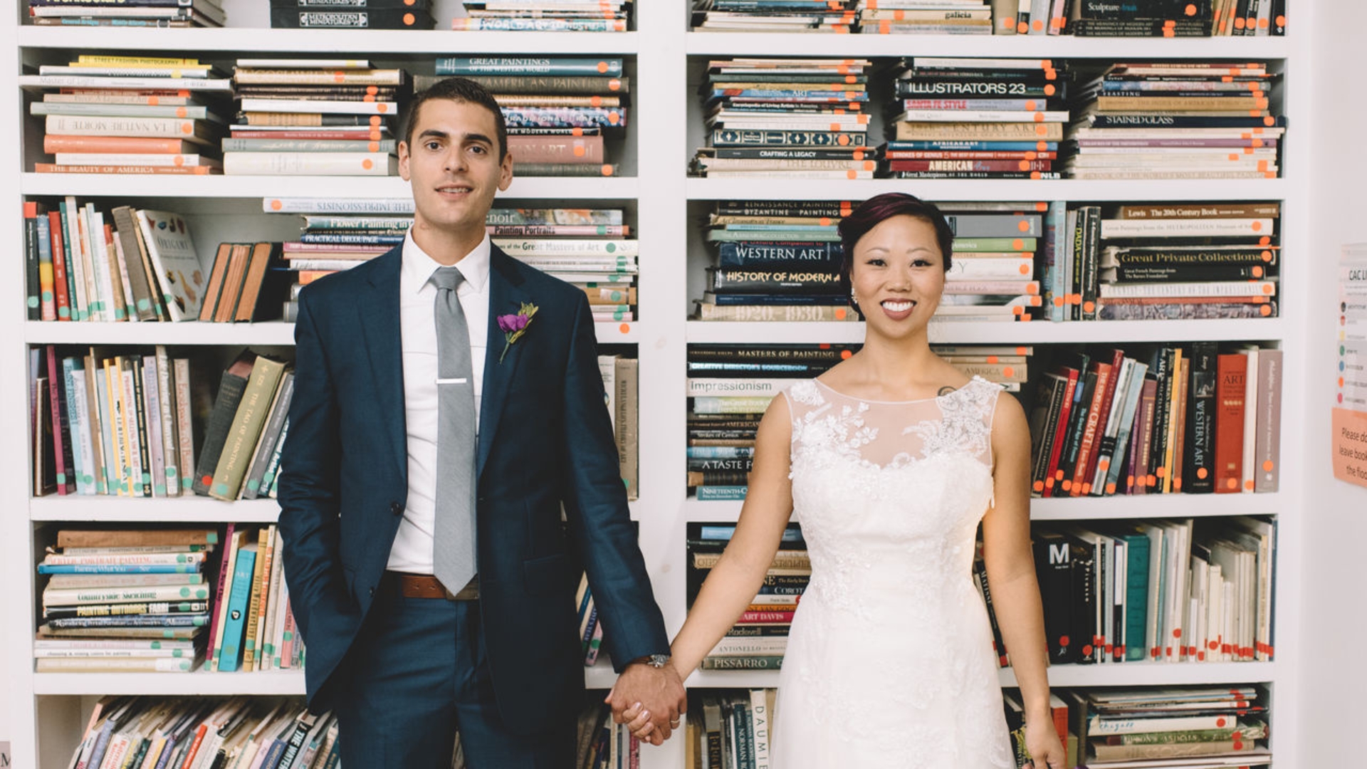 Bride and groom holding hands, standing in front of a bookshelf filled with books. The bride is in a white dress, and the groom is in a suit.