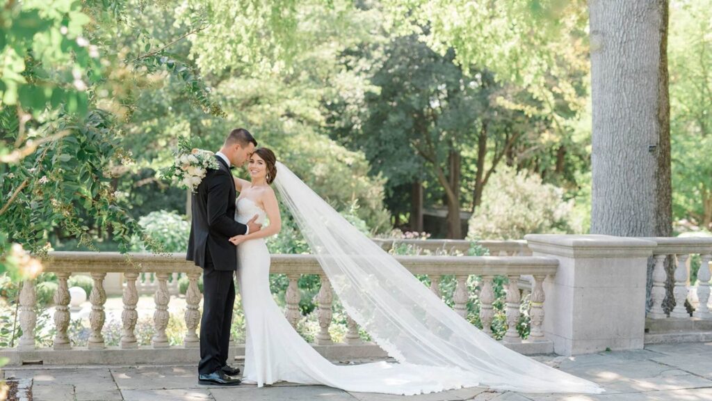 A bride and groom stand on a stone terrace surrounded by greenery. The bride wears a long veil, and the groom is in a black suit. They gaze at each other, holding a bouquet.