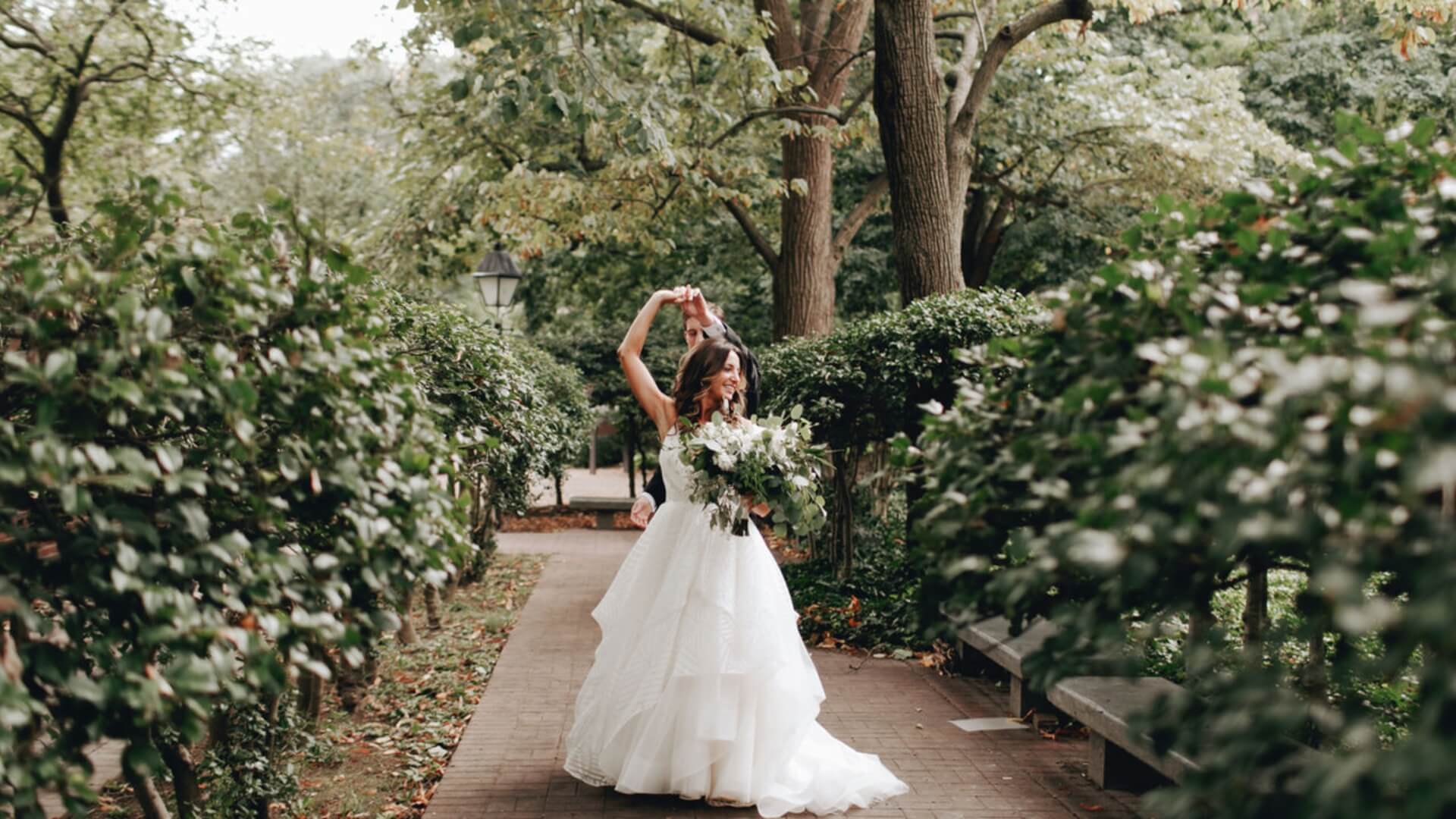 A bride in a white gown dances with a groom on a brick pathway surrounded by greenery in a garden. She holds a bouquet of flowers.
