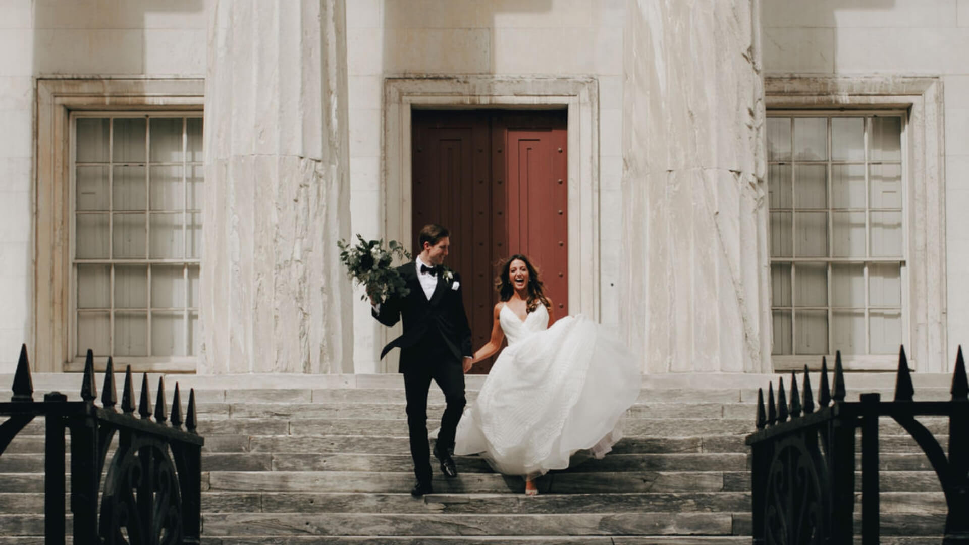 A bride and groom joyfully walk down marble steps, with the groom holding a bouquet.