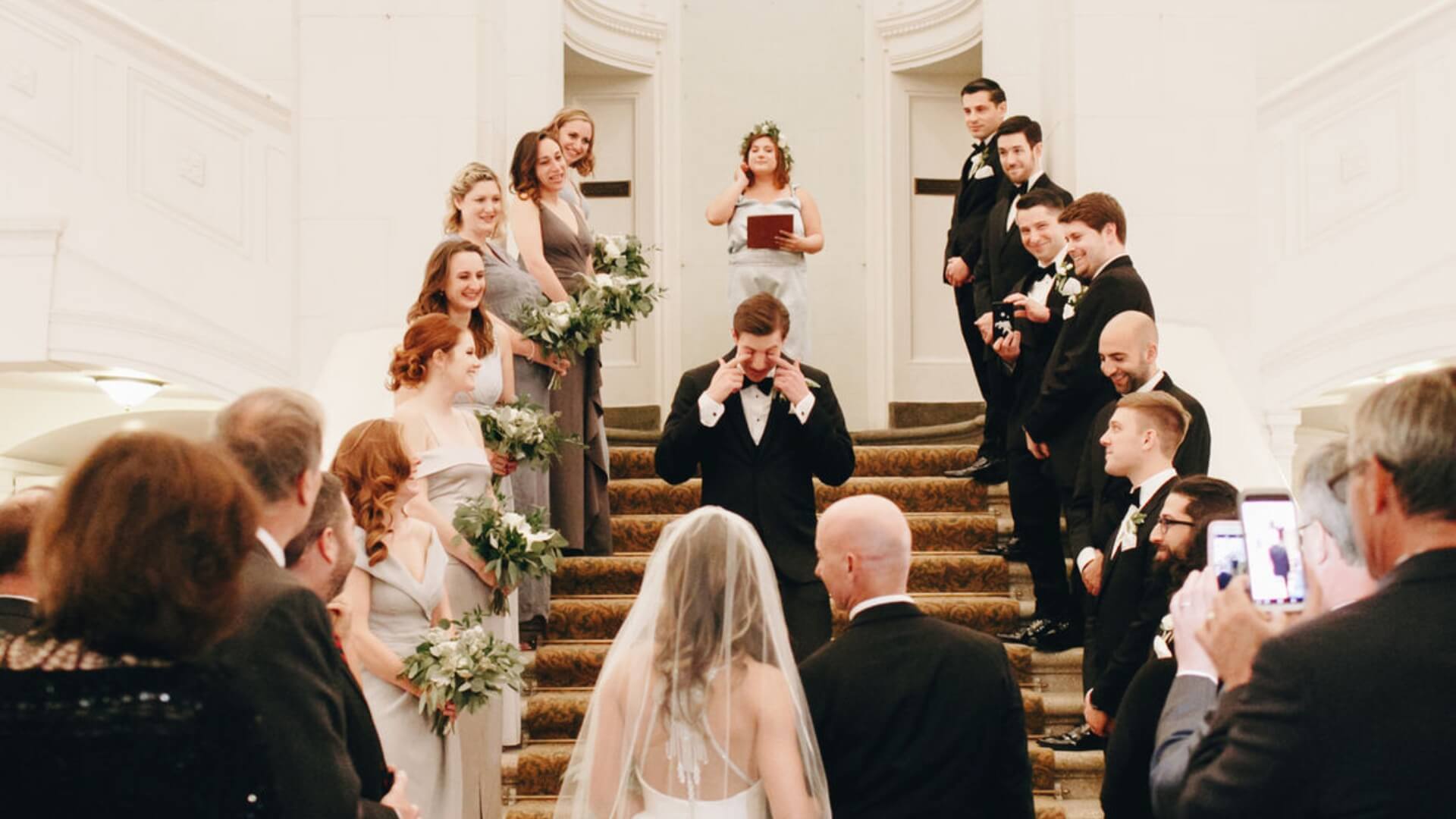 A wedding party poses on a staircase. The bride and groom are at the center, with bridesmaids and groomsmen on either side. Guests, some taking photos, are gathered below.