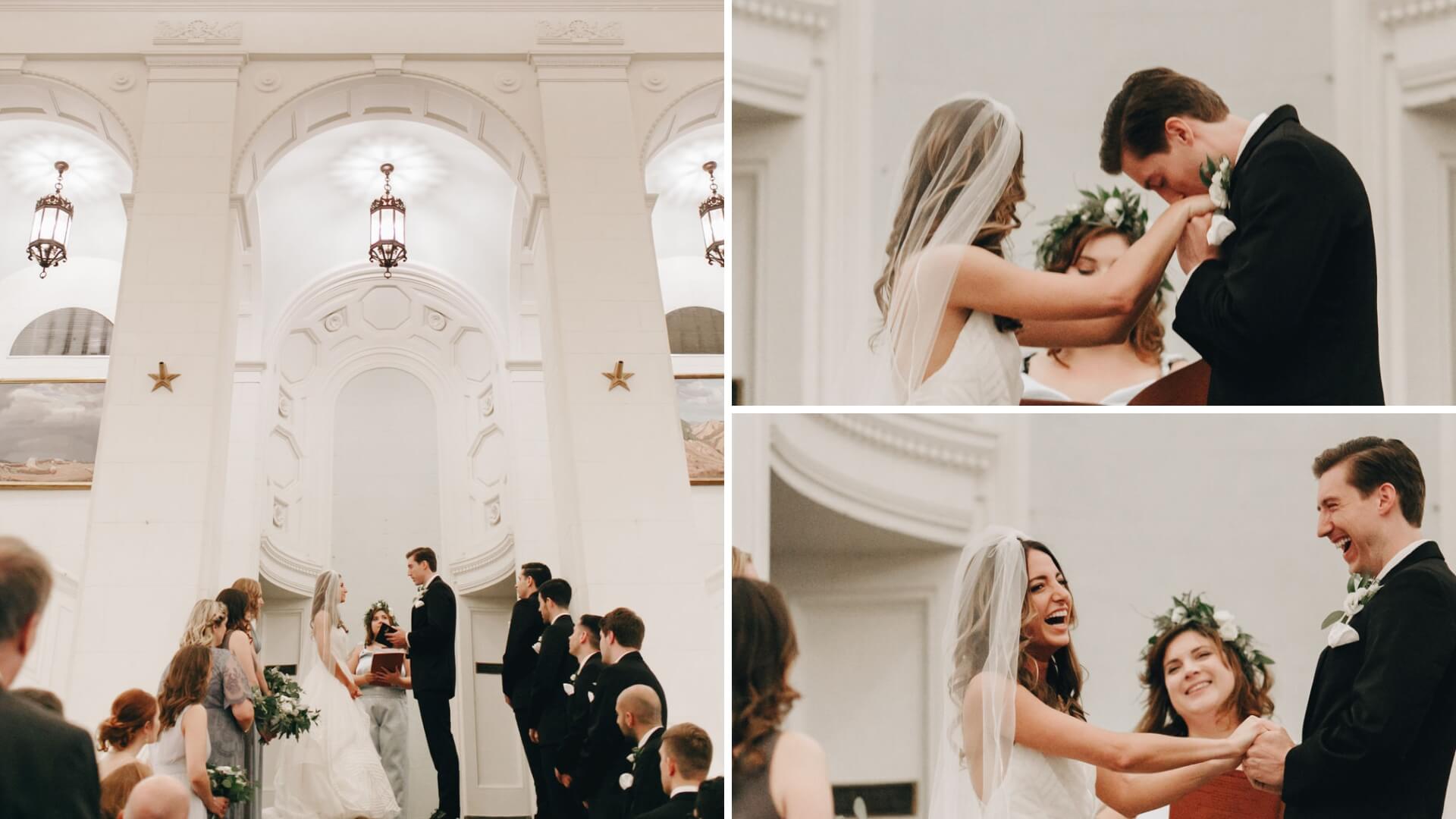 A bride and groom exchange vows and laugh during their wedding ceremony. They stand in a large ornate room with high ceilings and white walls. Bridesmaids and groomsmen surround them.