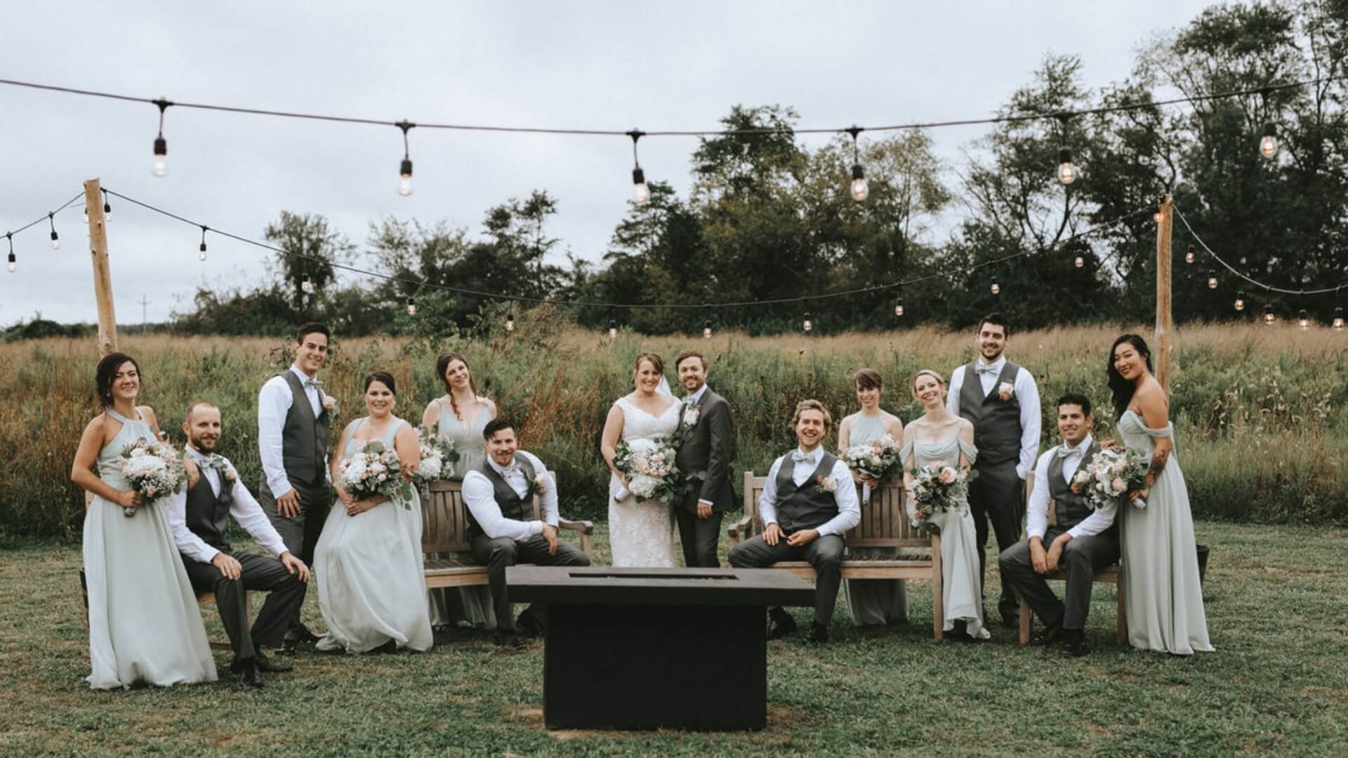 A wedding party poses outdoors. The group includes the bride, groom, bridesmaids, and groomsmen dressed in formal attire, arranged on wooden benches with a field and string lights in the background.
