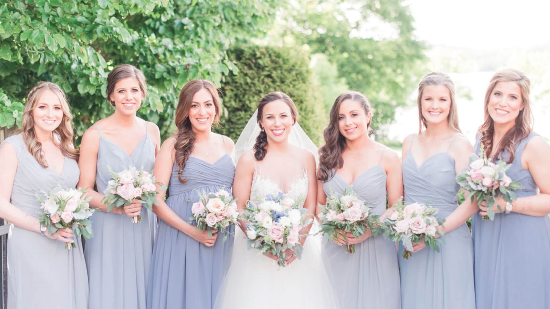 A bride stands with six bridesmaids outdoors, all wearing light blue dresses and holding bouquets. Green foliage is in the background.