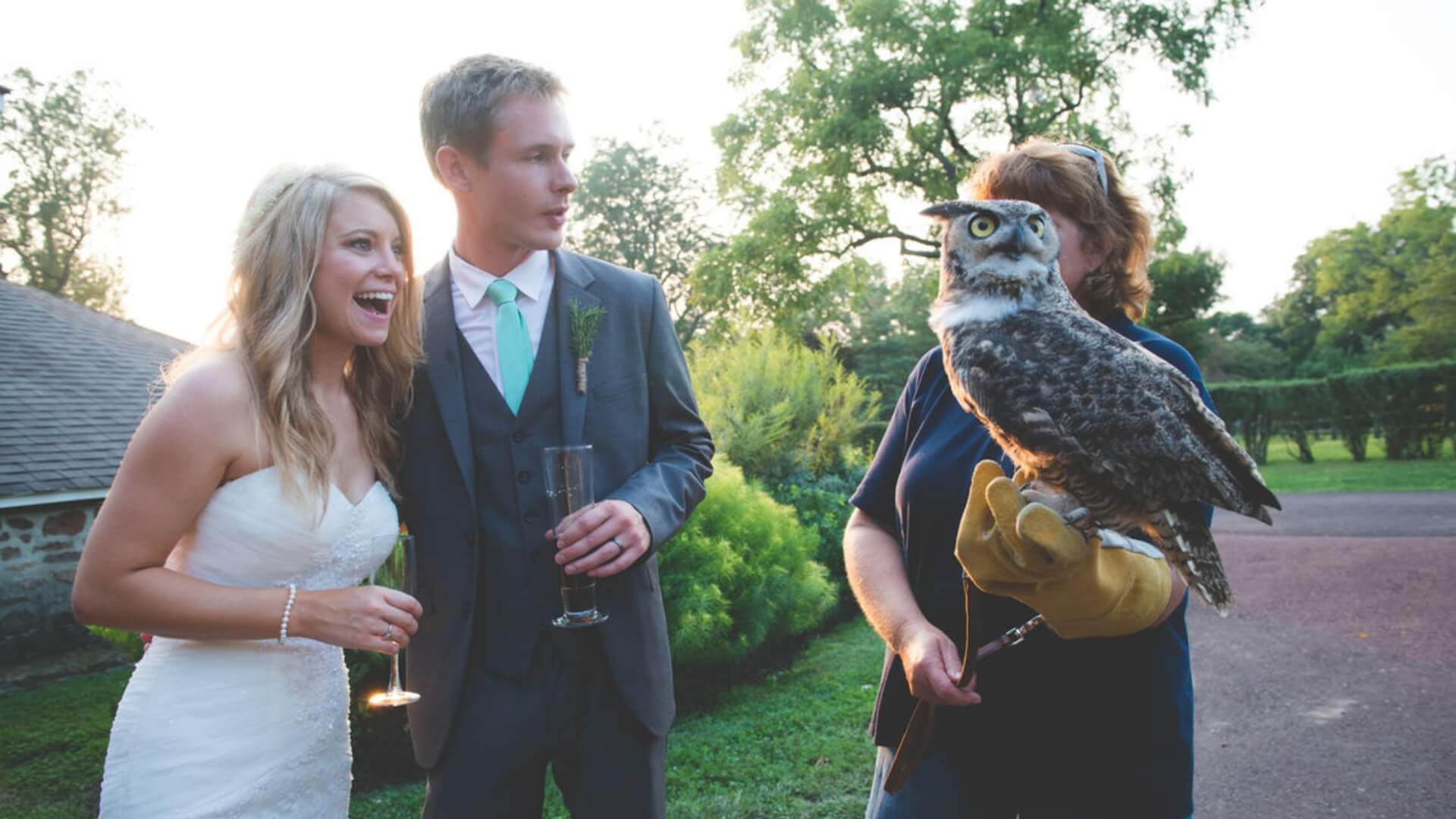 Bride and groom stand outdoors, smiling at a person holding a large owl on their gloved hand. Trees and a building are in the background.