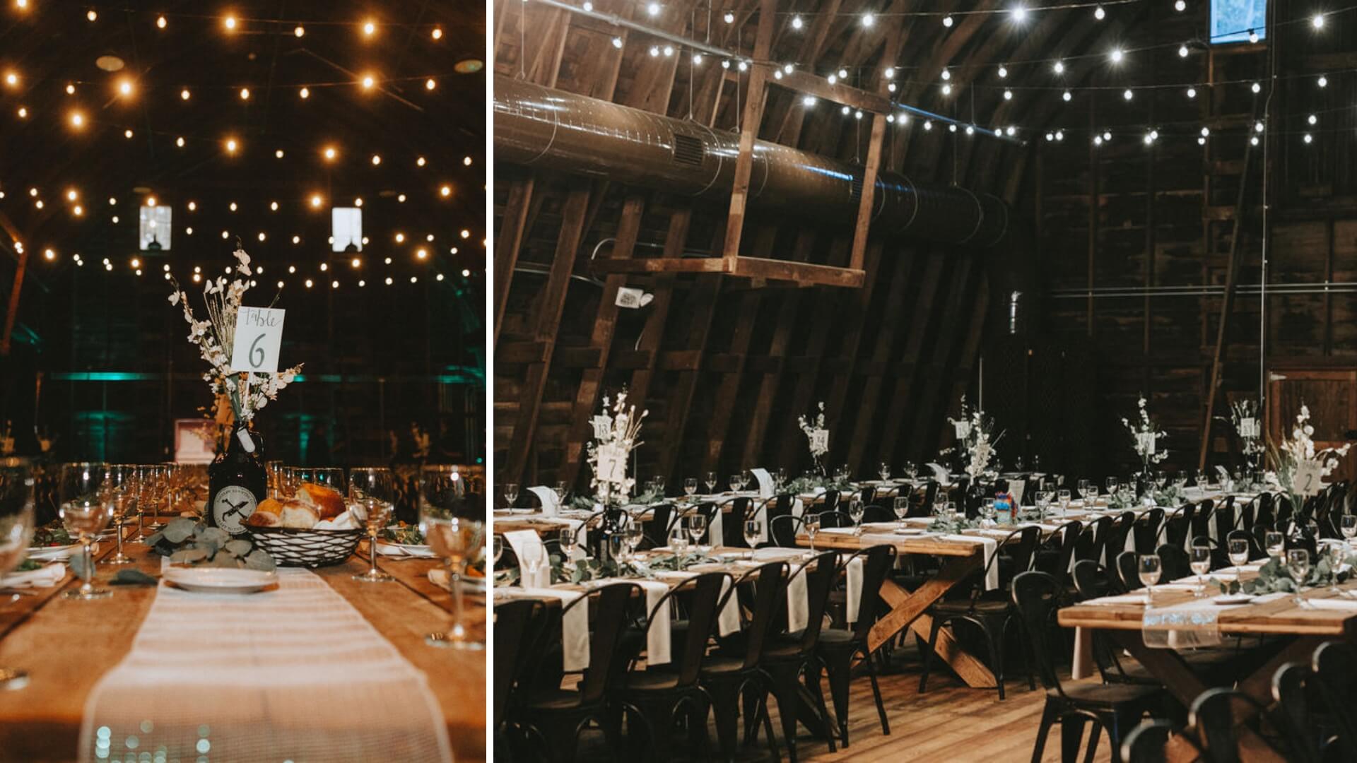 Rustic indoor wedding reception with long wooden tables, white flower centerpieces, and string lights overhead. Tables are set with plates, glasses, and table number cards.